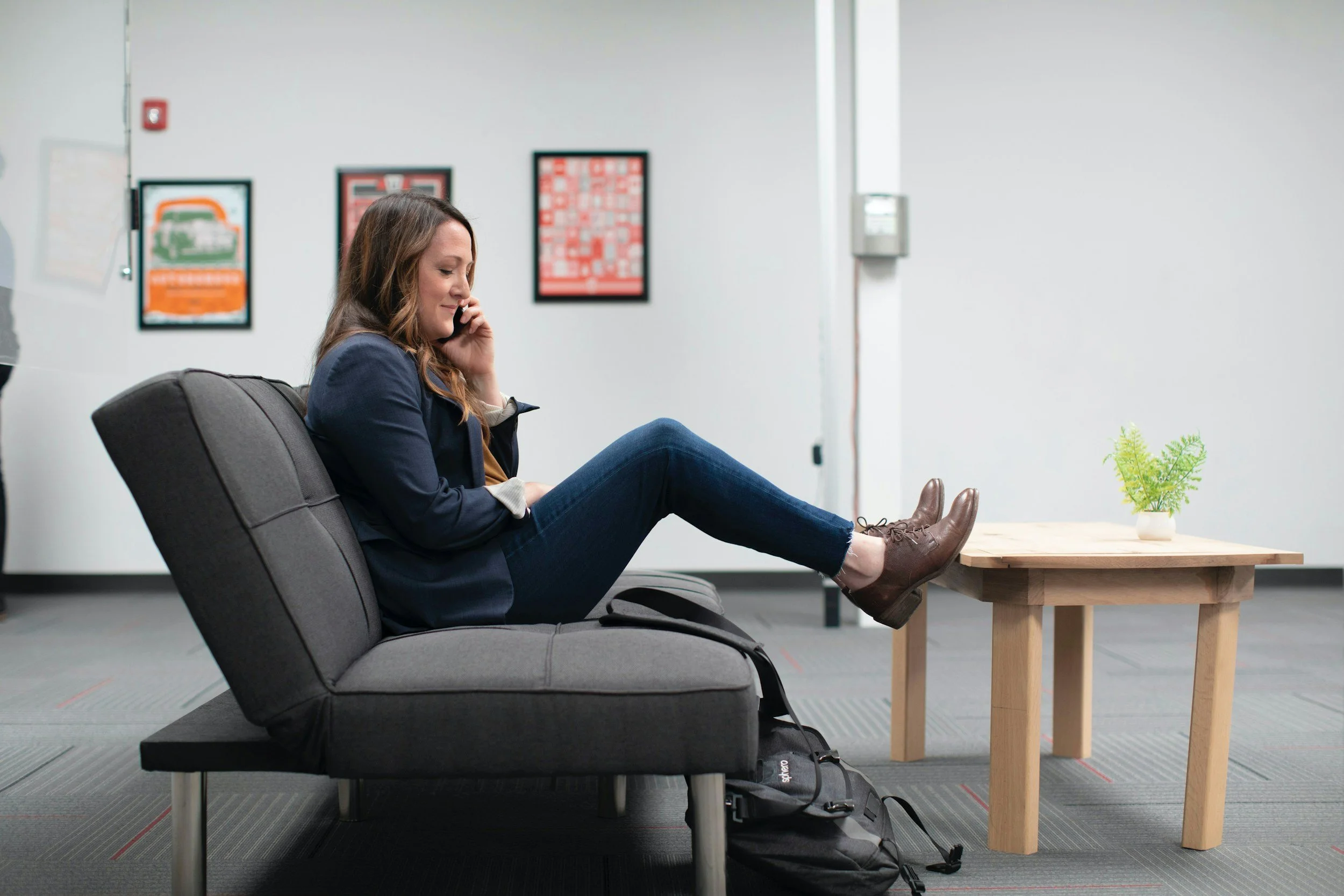 Woman sitting on a gray couch in a waiting area, talking on her phone, with her feet resting on a wooden table, and a black backpack on the floor beside her.