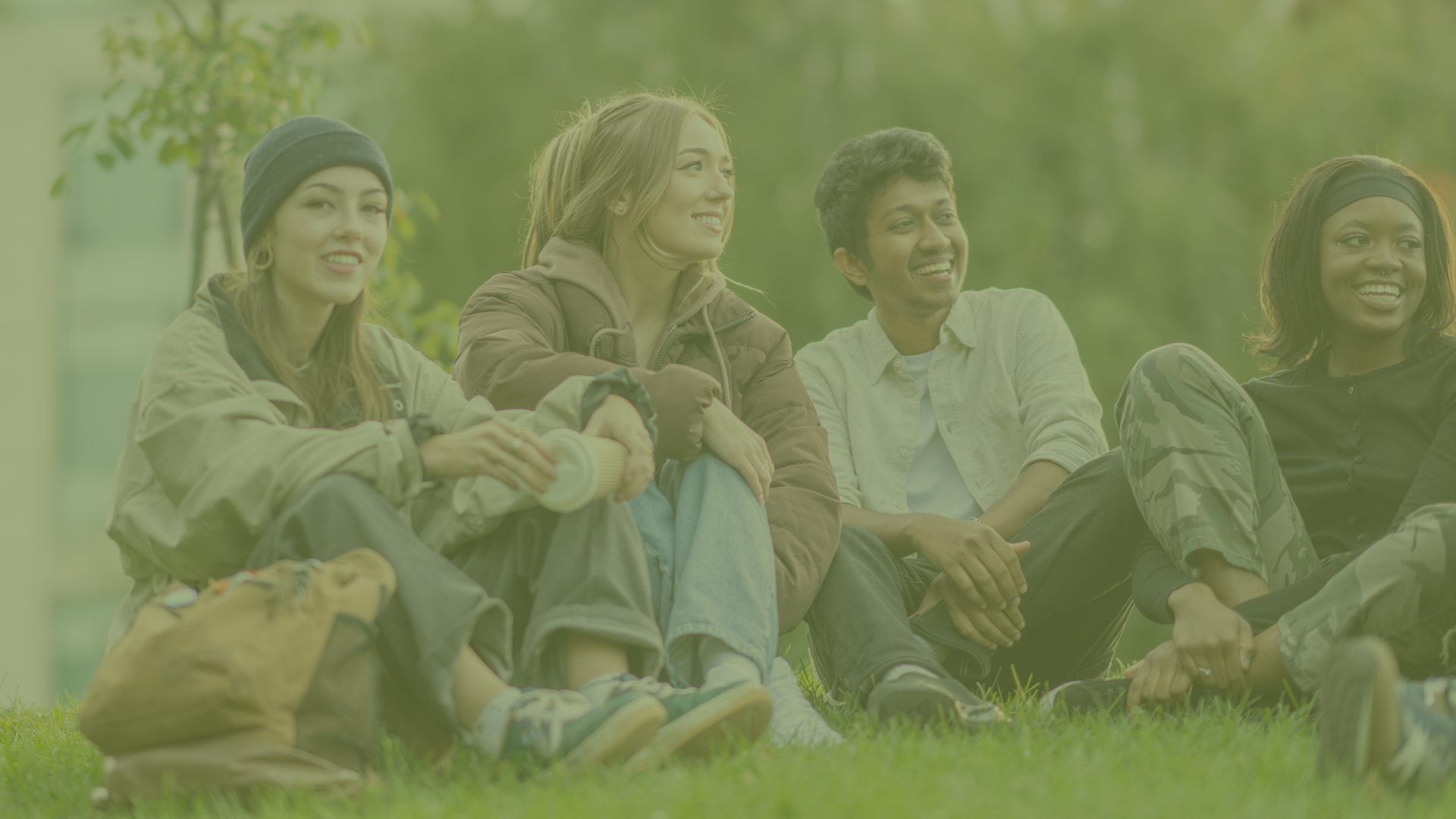 Four young people sitting on grass outdoors, smiling and talking in a park with trees in the background.