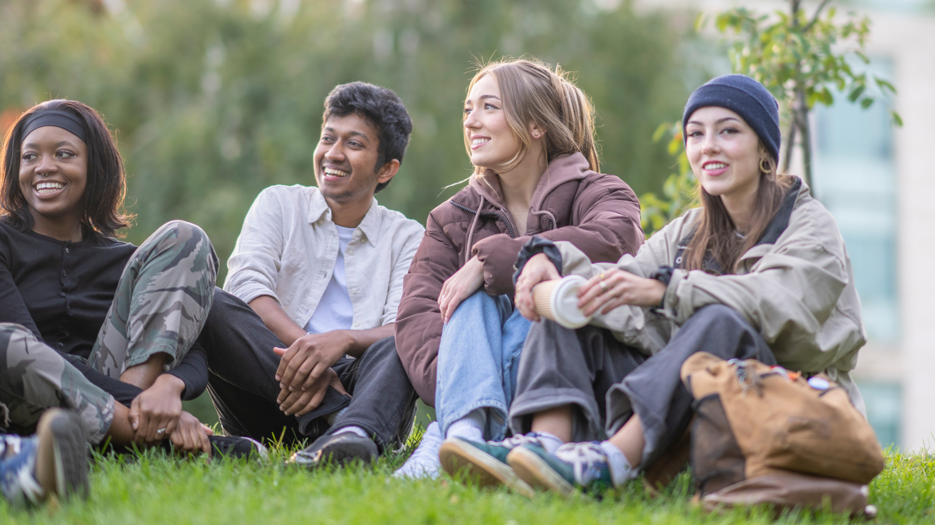 Four young friends sitting on grass in a park, smiling and chatting, with trees and a building in the background.