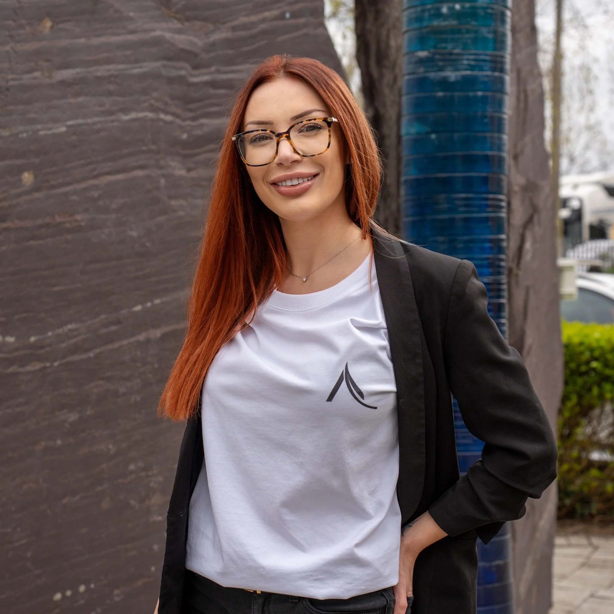 A woman with long red hair and glasses smiling outdoors, wearing a black blazer over a white T-shirt with a black graphic design, standing near a tree with a blue and black striped sculpture in the background.