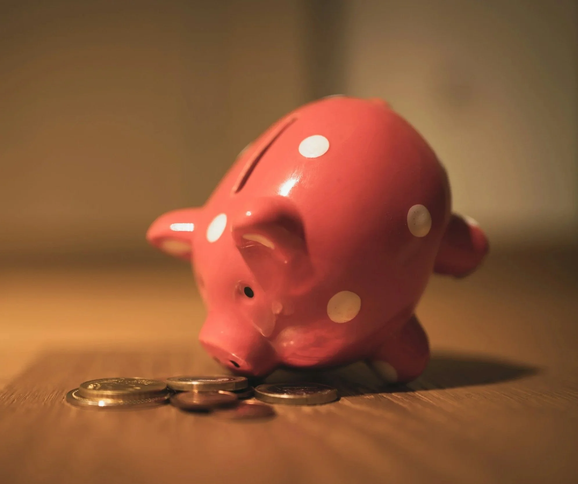 Pink piggy bank with white polka dots on a wooden surface, surrounded by coins, dimly lit.