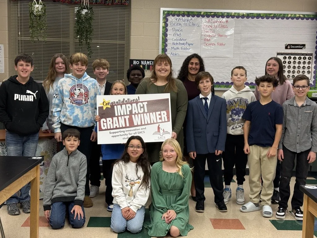 Group of children and two adults in a classroom holding a large sign that reads 'Congratulations! Impact Grant Winner' with a seal from SACS. The group is standing in front of a classroom whiteboard with educational posters and decorations.