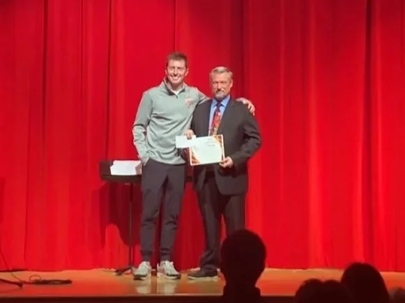 Two men stand on a stage with a red curtain in the background, one holding a certificate and the other arm around his shoulder.