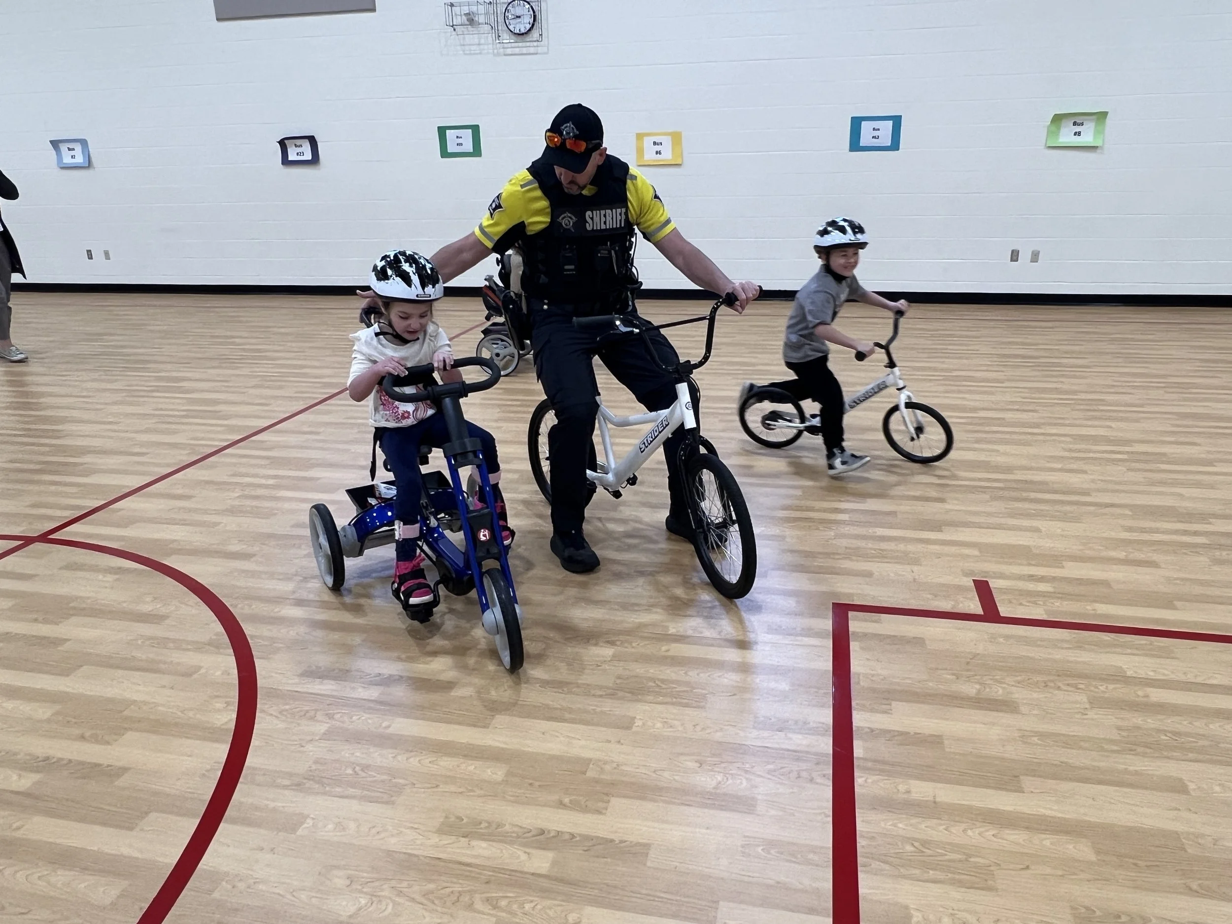 A police officer helping two young children learn to bike inside a gymnasium. The children are wearing helmets, and one child is riding a tricycle while the other is on a small bike. The gym has a wooden floor and colorful signs on the wall.
