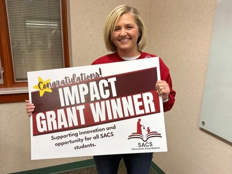 A woman with blonde hair smiling and holding a large sign that reads 'Congratulations! Impact Grant Winner' at an indoor event.