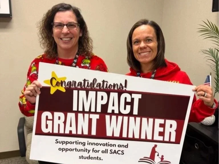 Two women smiling and holding a sign that says 'Congratulations! Impact Grant Winner Supporting innovation and opportunity for all SACS students' in an indoor setting.