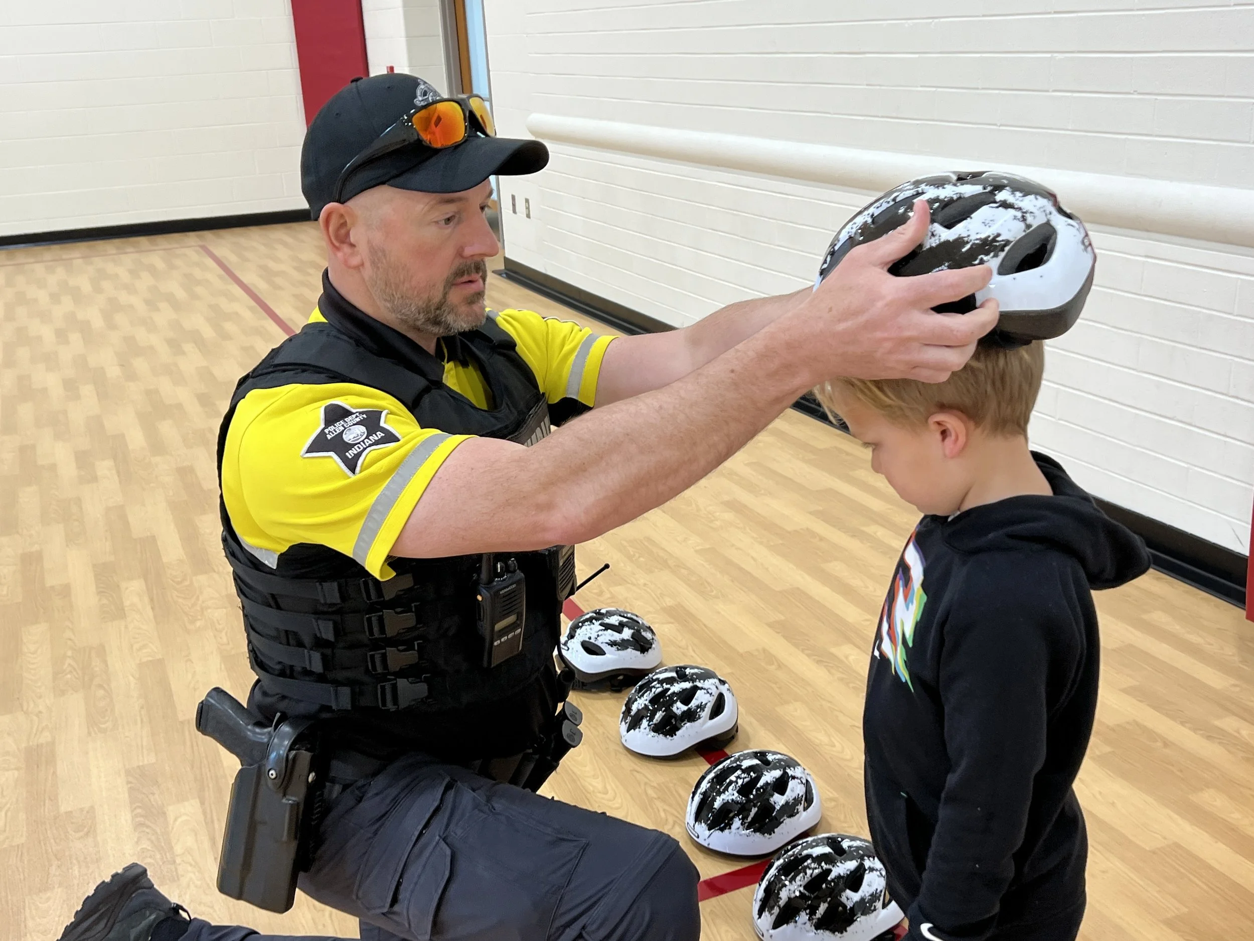 A police officer fitting a black and white bicycle helmet on a young boy's head inside a gymnasium, with several other helmets on the floor nearby.