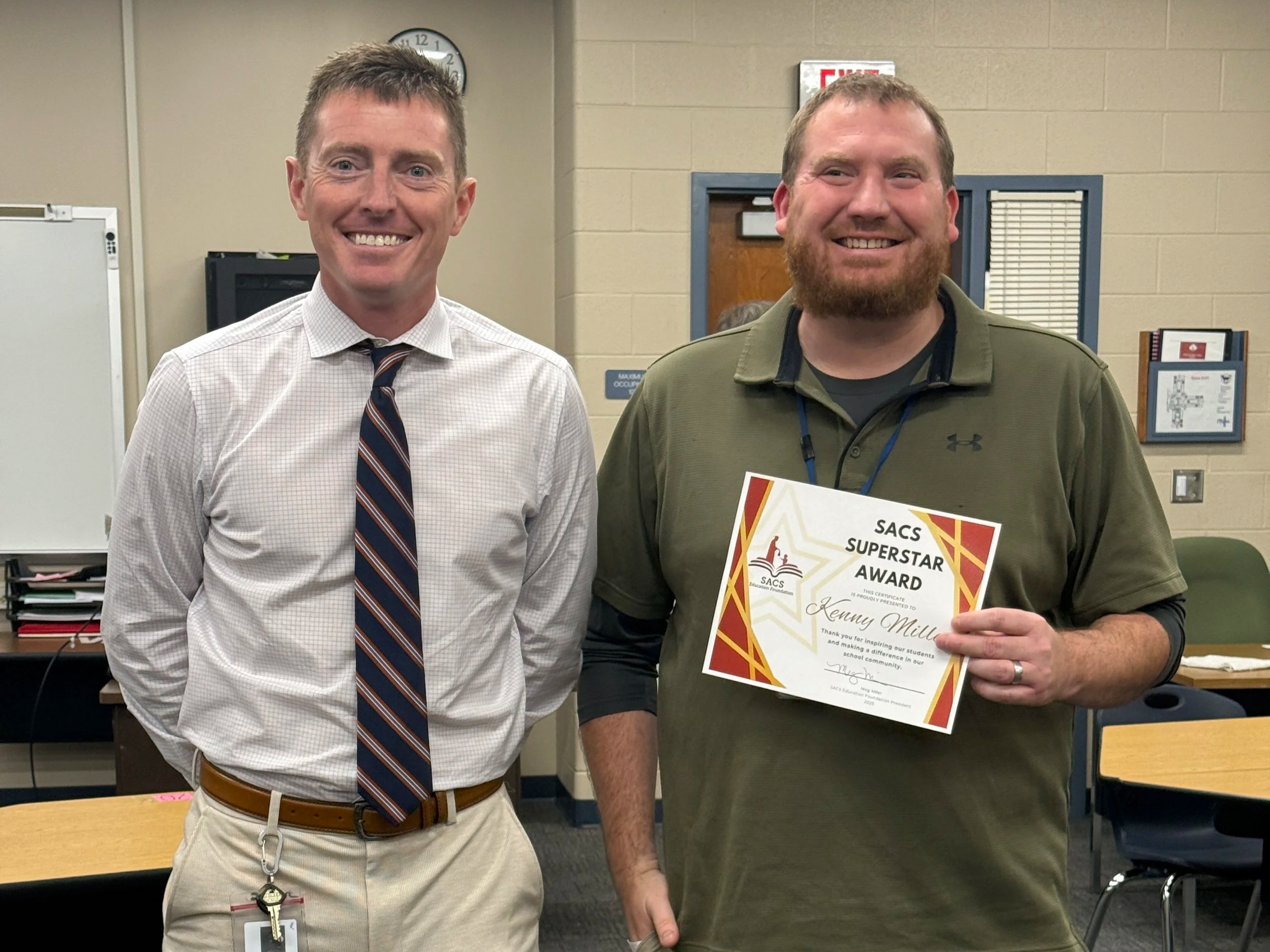 Two men standing together in a classroom or meeting room, smiling. One is wearing a white shirt and striped tie, the other is wearing a green polo shirt and holding a certificate that reads 'SACS Superstar Award'.