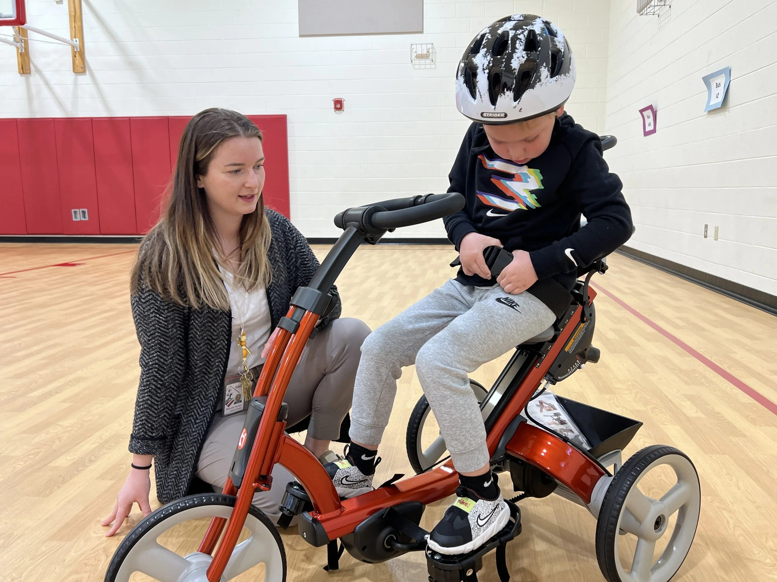A young boy wearing a helmet is sitting on a red adaptive tricycle, with a woman kneeling beside him, assisting him as he adjusts the seat belt in a gymnasium.
