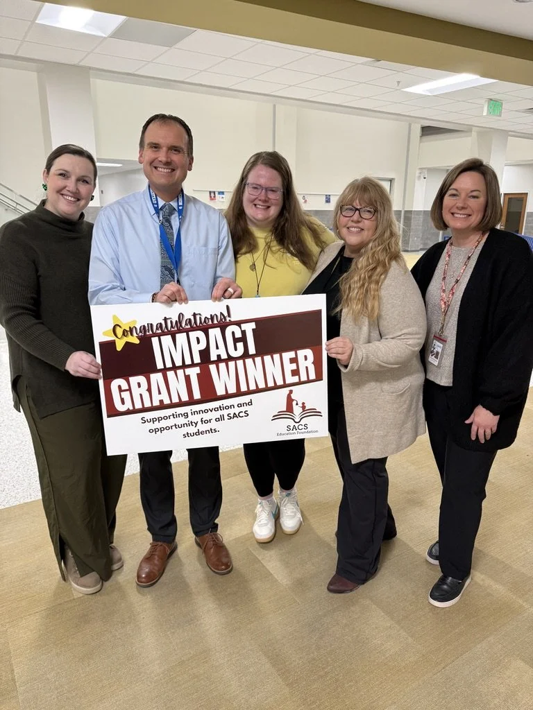 Group of five adults smiling, holding a sign that says 'Congratulations! Impact Grant Winner' at an indoor event.