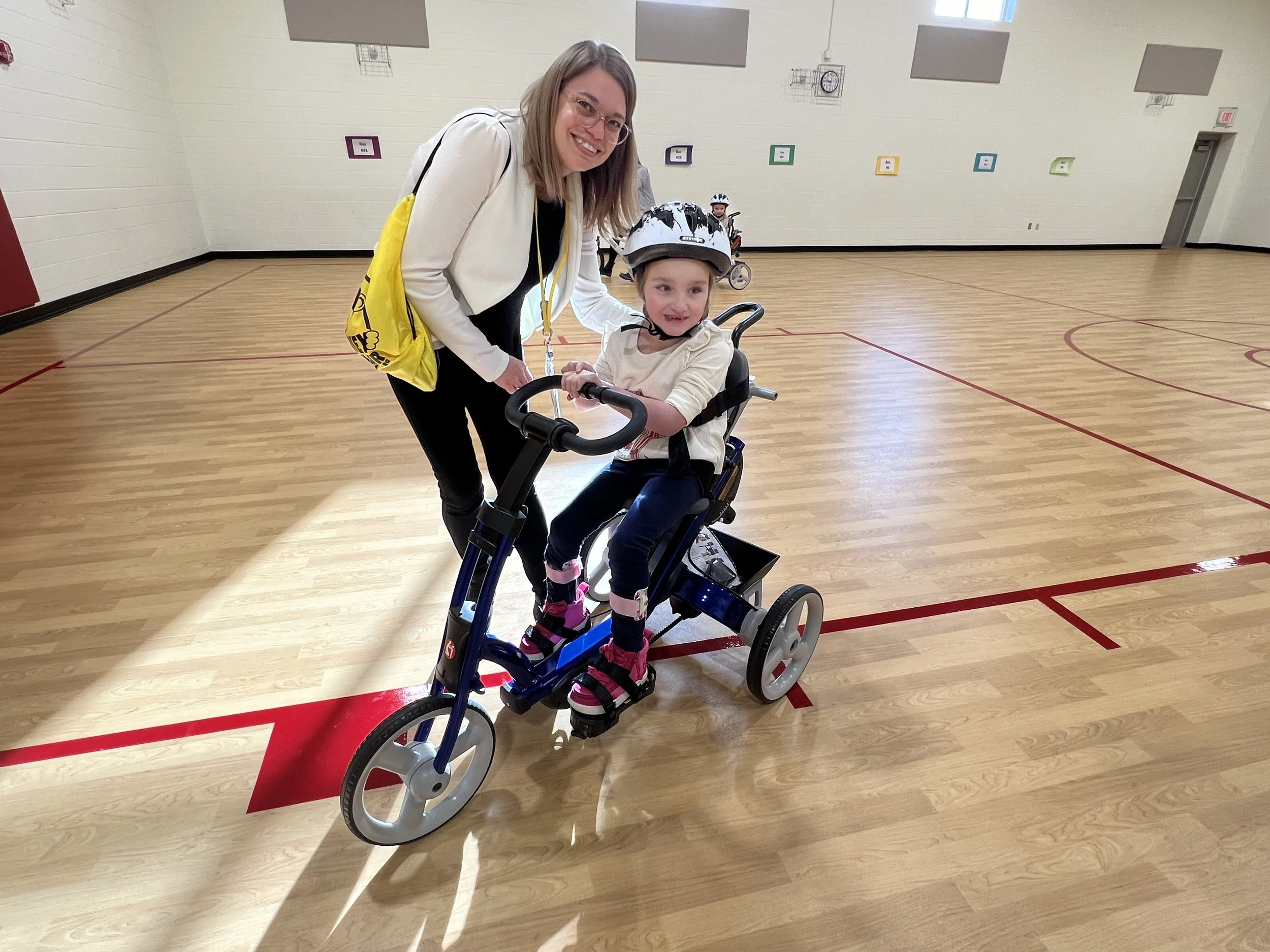 A smiling woman helping a young girl on a tricycle in an indoor gym with a wooden floor and colorful wall decorations.