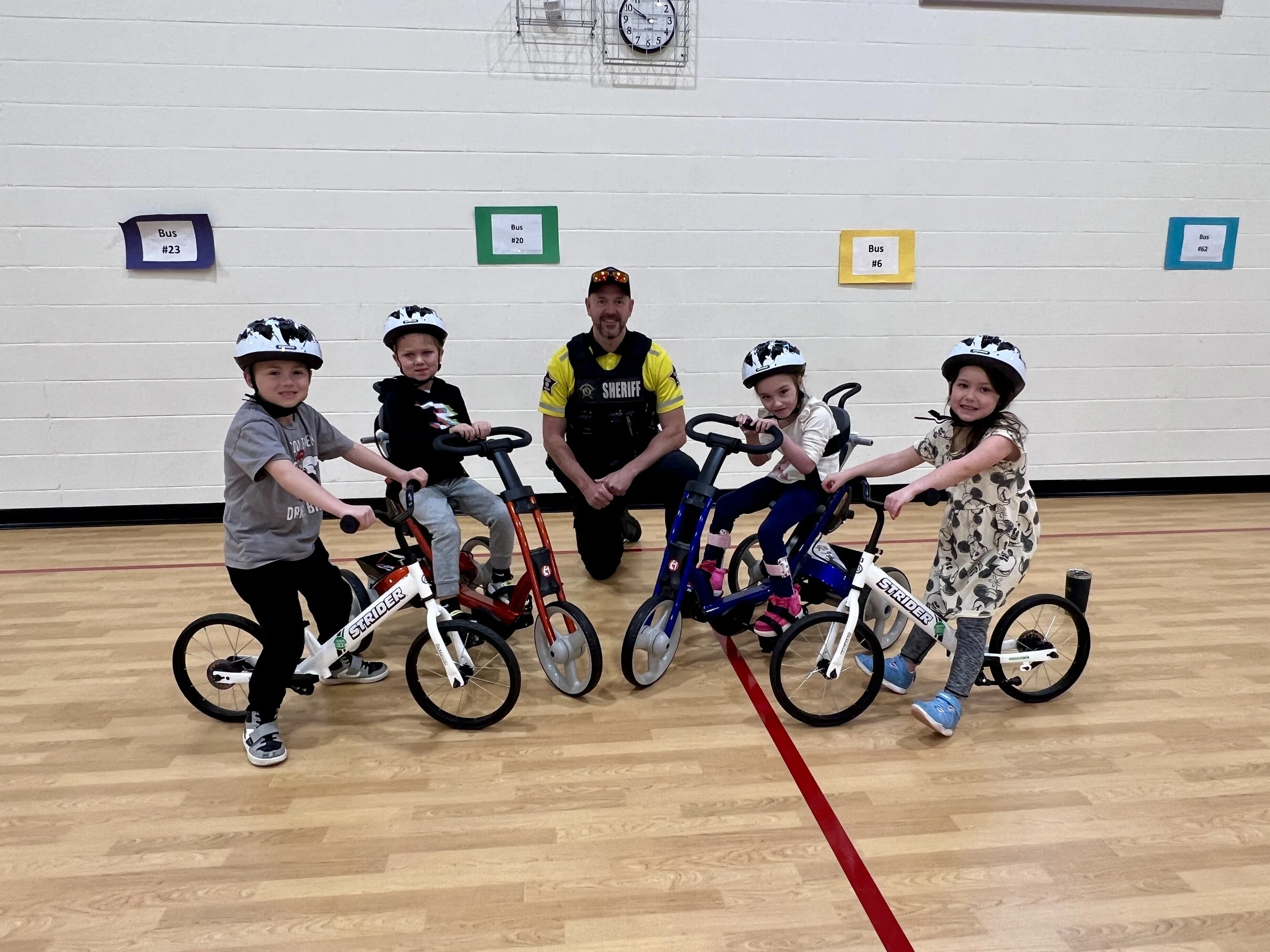 A group of four young children and a police officer pose with bikes inside a gymnasium, all wearing helmets.
