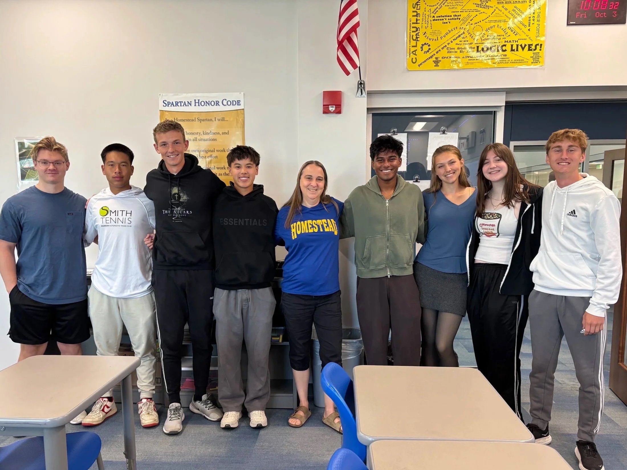 Group of nine students and a teacher standing in a classroom, posing for a photo with a white wall, posters, and furniture in the background.