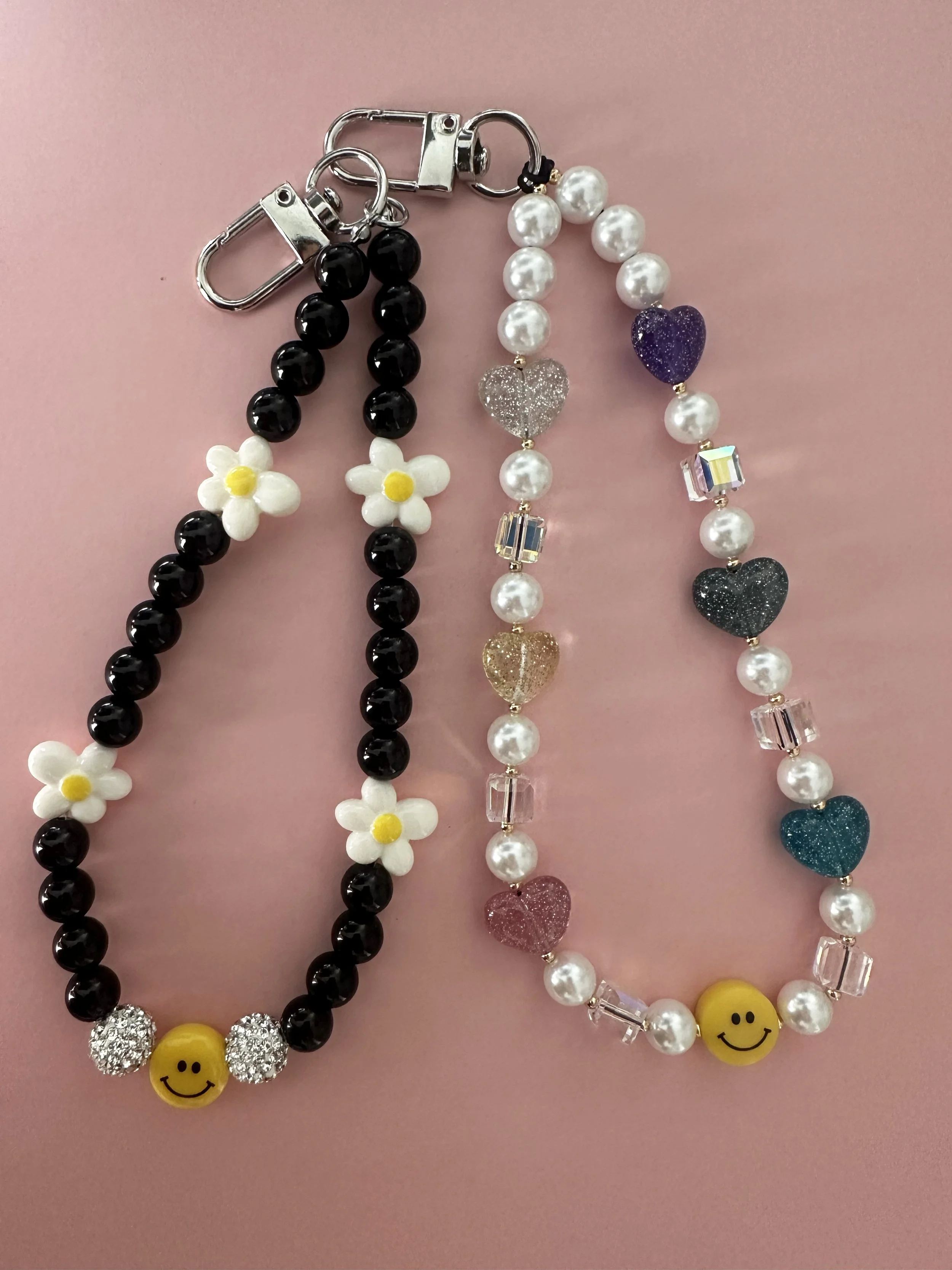 Two beaded bracelets on a pink background. The bracelet on the left has black beads with three white daisy flower beads and a yellow smiley face bead. The bracelet on the right has white beads with colorful glittery heart beads, clear rectangular beads, and a yellow smiley face bead.
