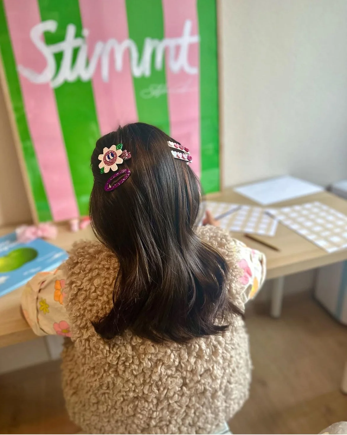 Back view of a young girl with long, dark hair decorated with colorful hair clips, sitting at a table with stationery and patterned paper, in front of a colorful background with the word 'Strawberry'.