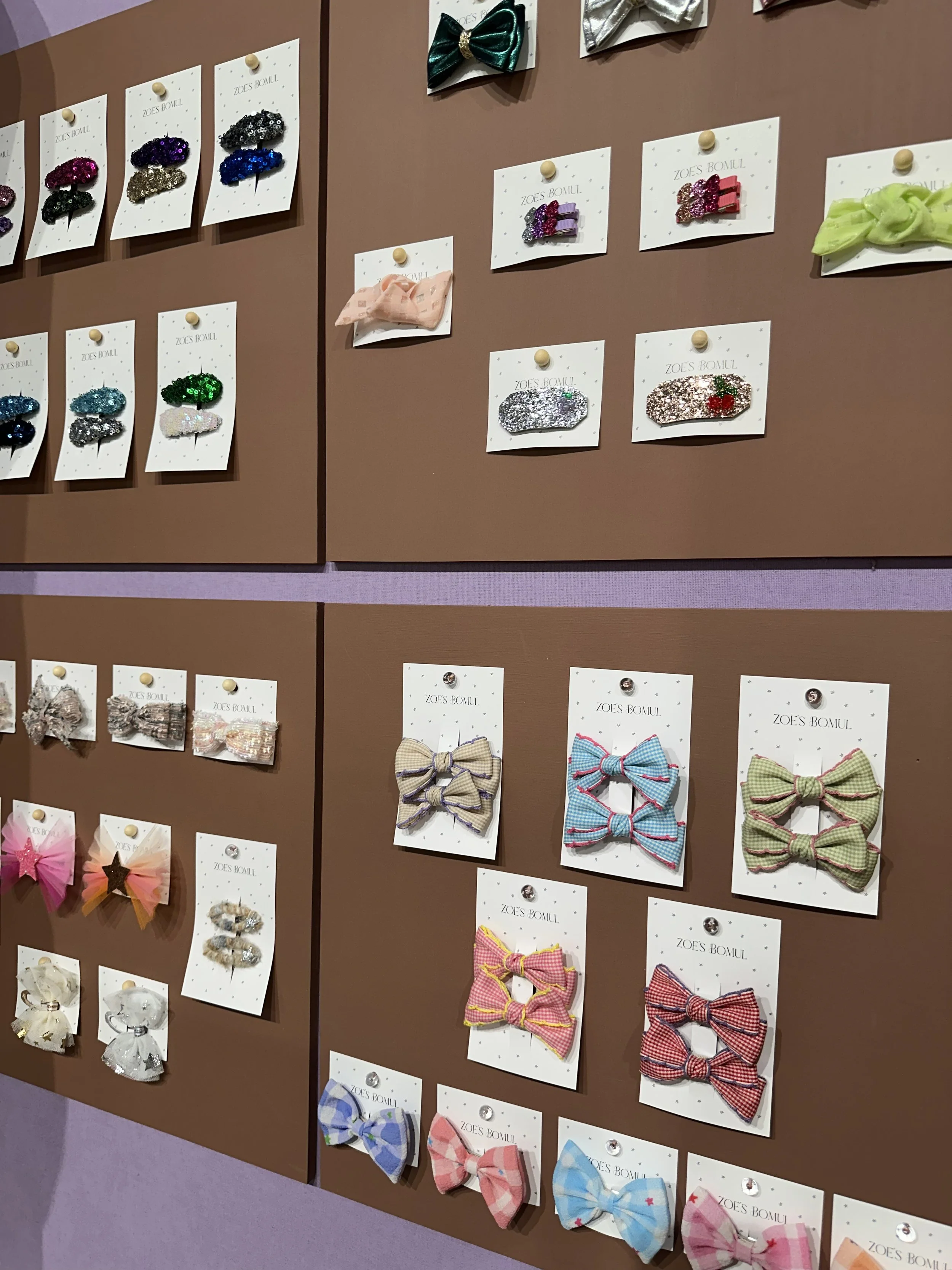 Display of various colorful hair accessories, including glittery hair clips, bows, and scrunchies, arranged on a brown board with white cards labeled 'Zoe's Bowlu'.