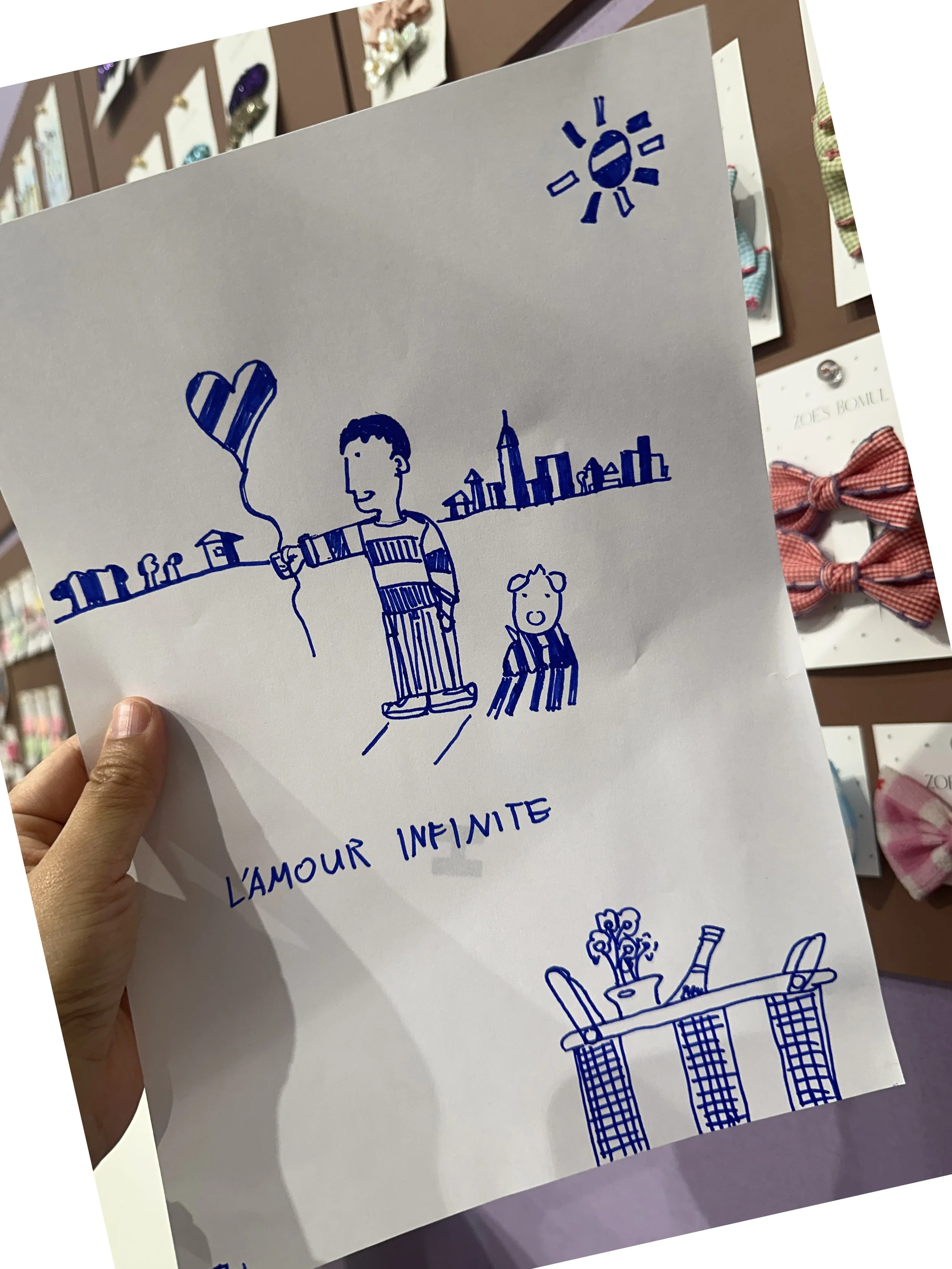 A hand holding a white paper with blue ink drawings of a person holding a heart-shaped balloon, a smaller child figure, cityscape background, a sun, and a table with flowers and a bottle. The text "L'AMOUR INFINITE" is written below the drawings.