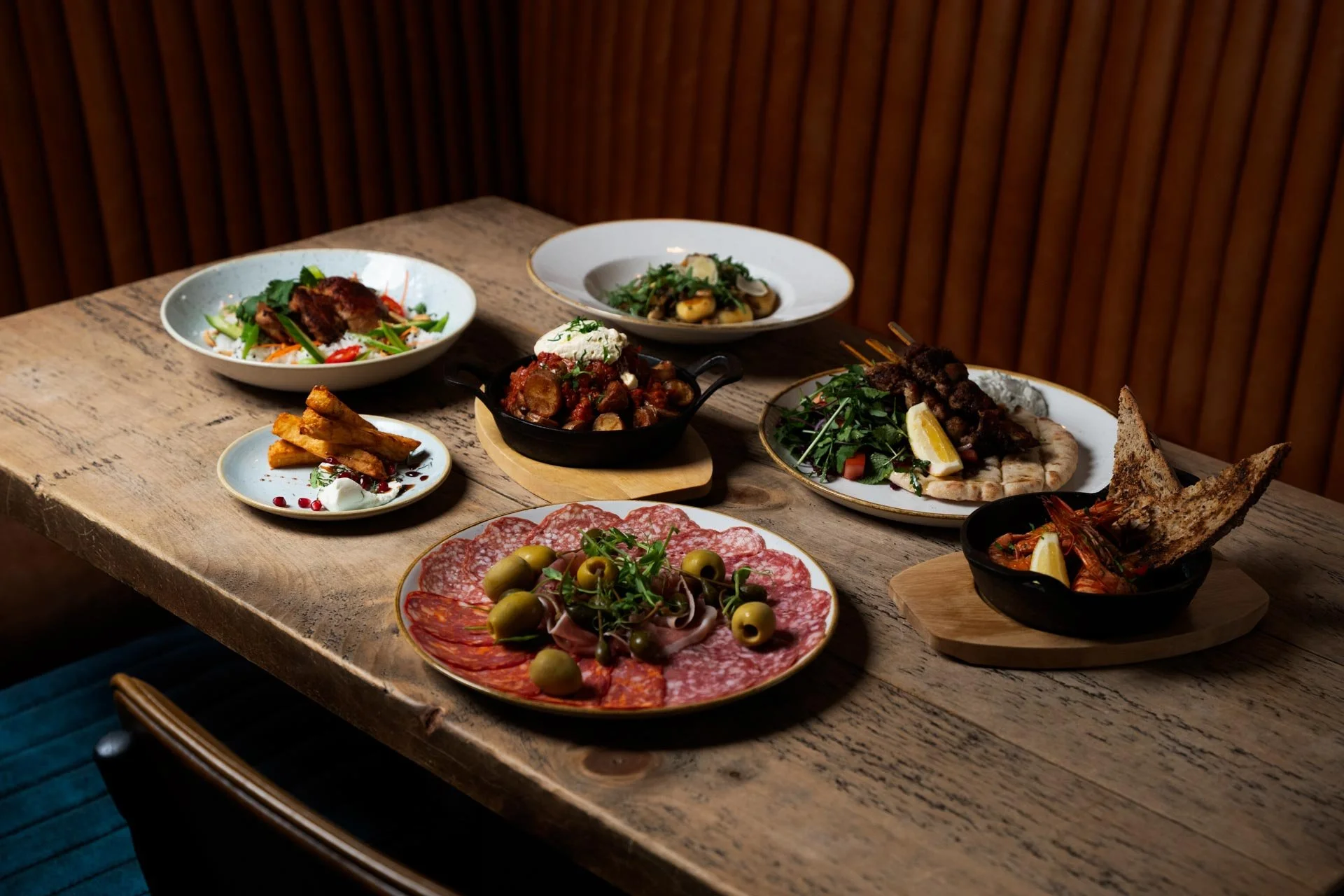 A wooden table with various plates of assorted appetizers and dishes, including charcuterie with olives, grilled meats, vegetables, and bread with prawns, pasta, pizza.
