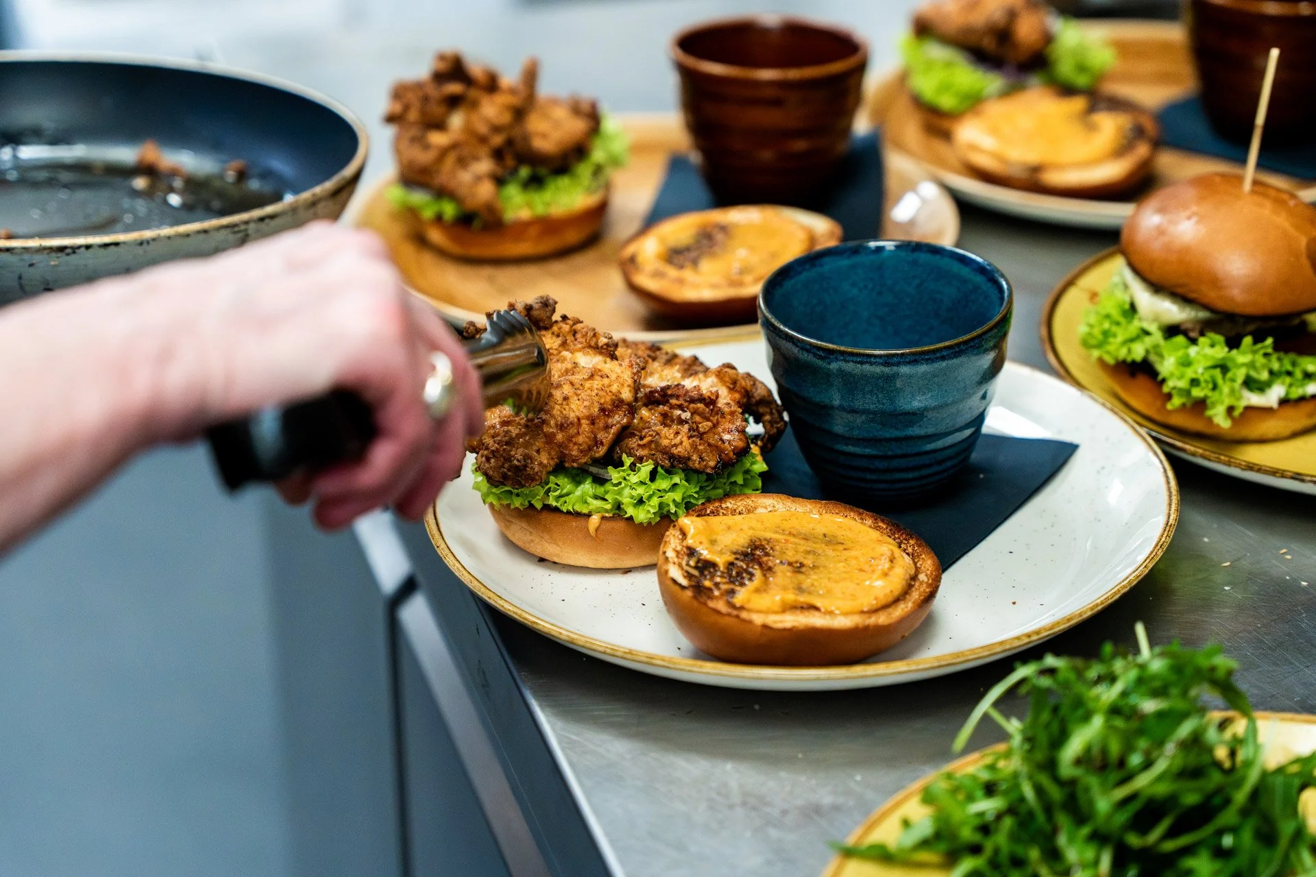Person placing a fried chicken burger onto a large white tray with various sliders and bowls of condiments and toppings on a kitchen counter. Perfect for catering.
