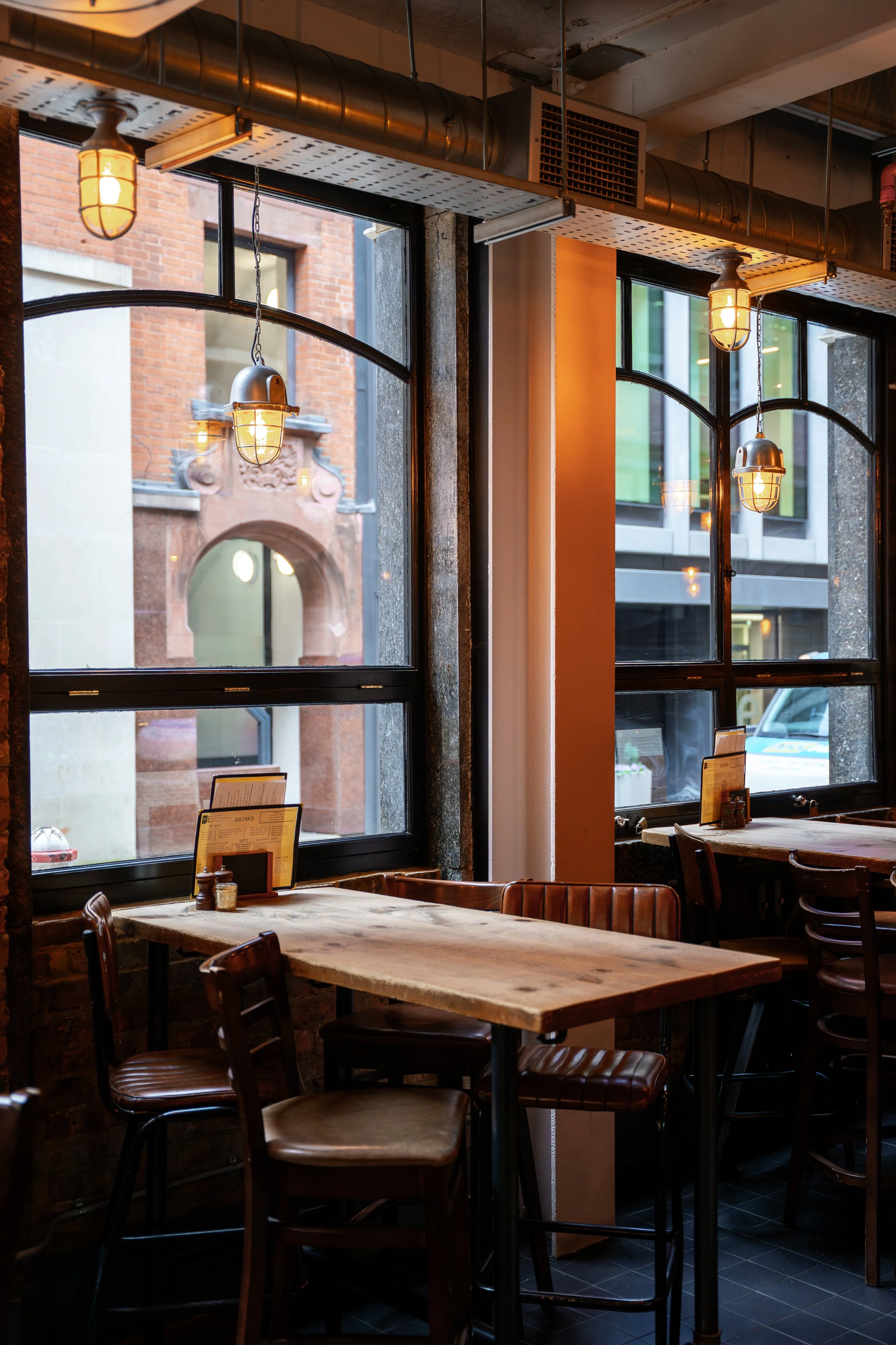 Interior of a restaurant and pub with large windows, wooden tables, leather chairs, exposed brick wall, and industrial lighting fixtures.