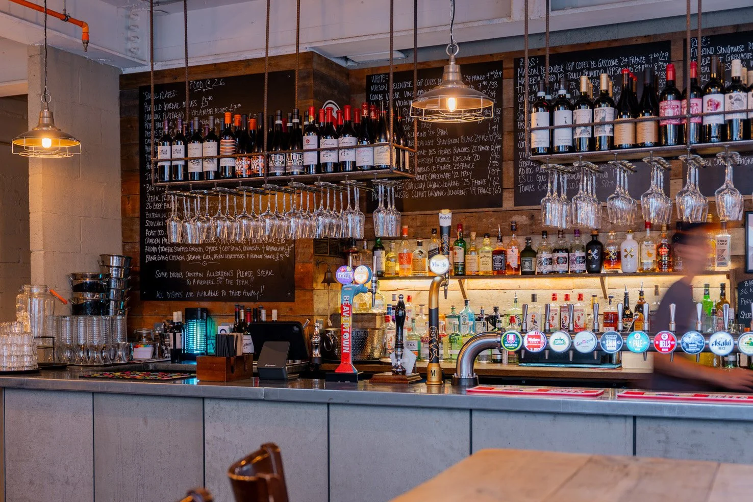 A chic, cool, industrial pub bar with hanging wine glasses, bottles of wine on shelves, and tap handles for draft beers. Chalkboard menus with handwritten text are behind the bar.