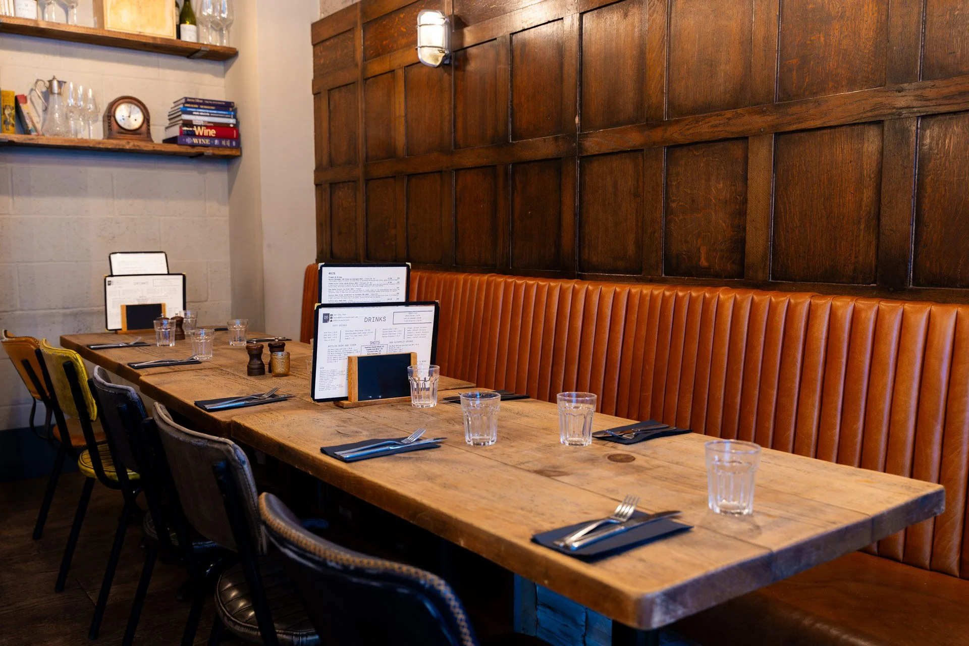 Interior of a cozy restaurant and pub with a long wooden table set with glasses, utensils, and menus. There is a high wooden panel on the wall and a leather-upholstered bench along the side. The ambiance features warm lighting and rustic decor.