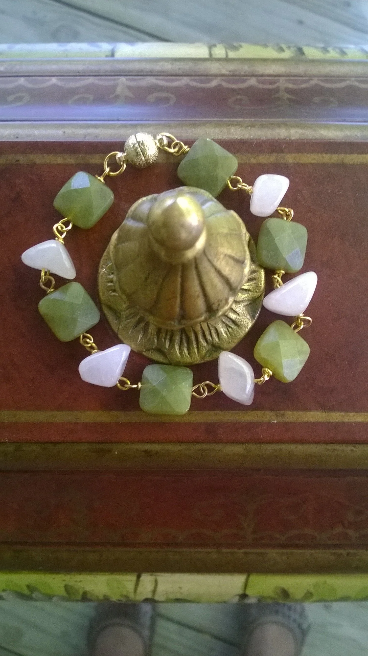 A gold-colored decorative object resembling a bell with a textured surface, placed on a wooden surface with a jewelry bracelet surrounding it. The bracelet has alternating green square beads and white triangular beads, connected by gold links.
