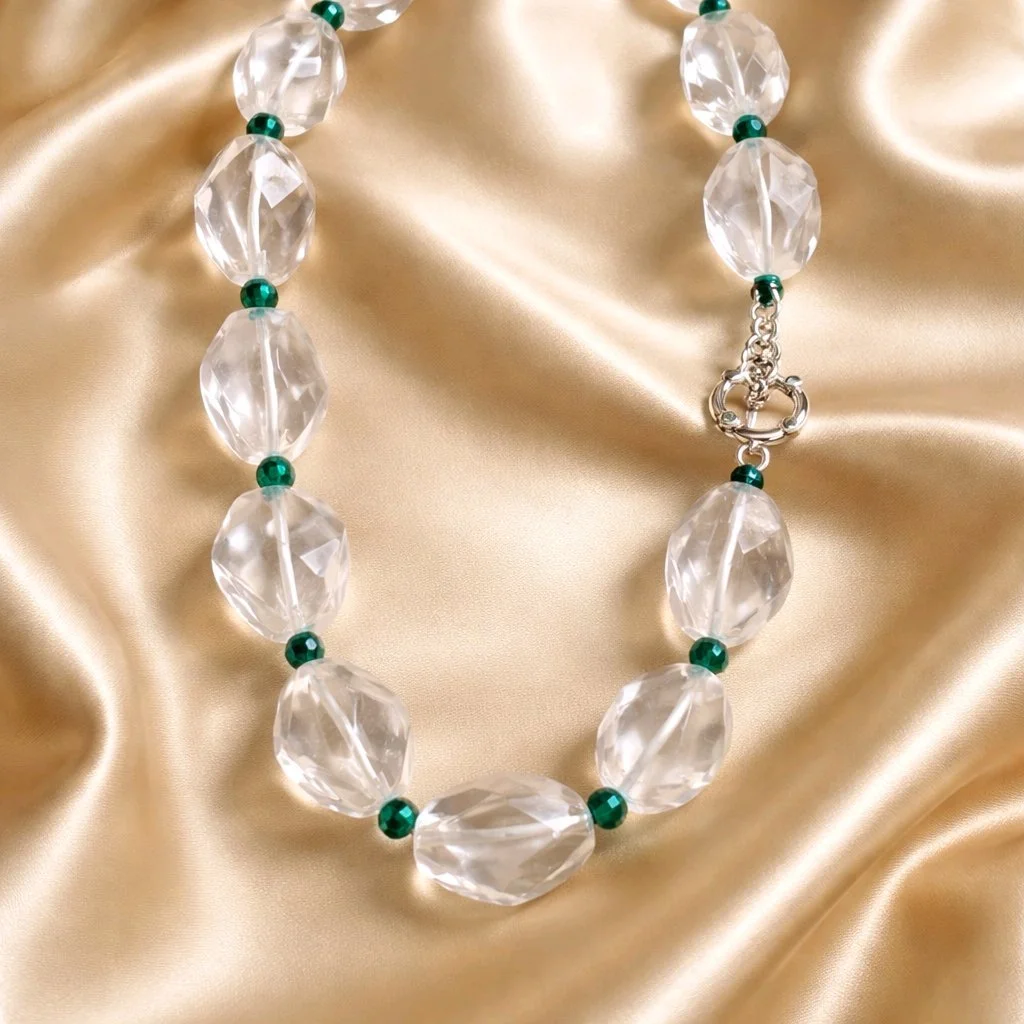 A necklace with large, clear, faceted beads alternating with small green beads, resting on a silky, gold-colored fabric.