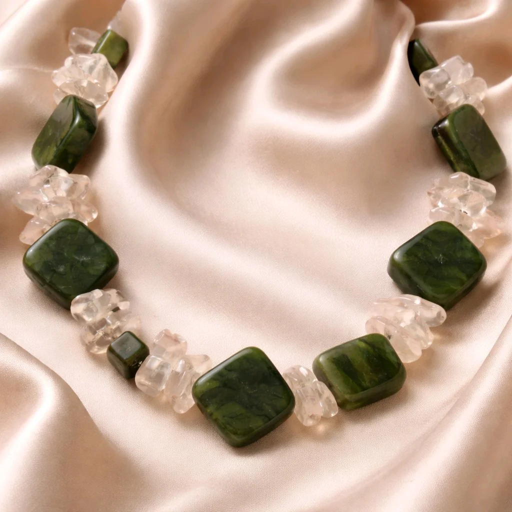 Necklace with large green square beads and smaller clear beads, displayed on shiny satin fabric.