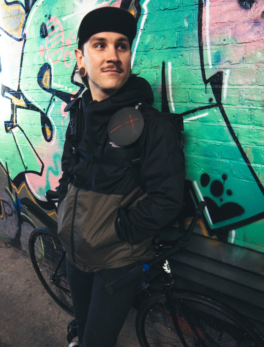A man with black hair, wearing a black cap, black jacket, and jeans, stands next to a black bicycle with a colorful graffiti wall behind him.
