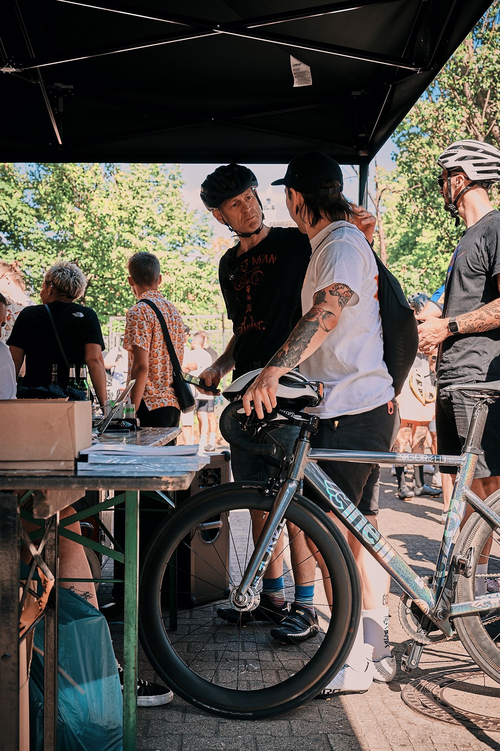 People gathered under a tent at an outdoor event, with most wearing helmets, some with tattoos, and a bicycle in the foreground.