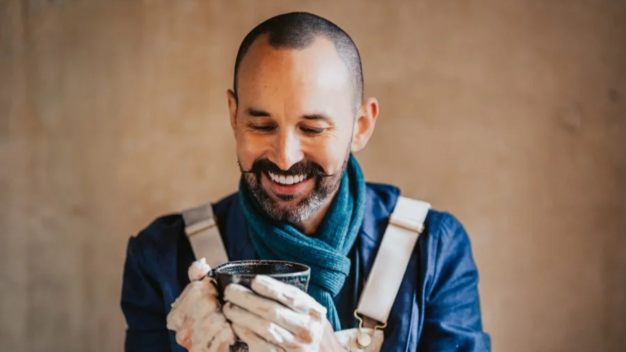 Images shows ceramic artist Jez Rose, founder of FOUND ceramics studio in Rutland holding a mug with porcelain covered hands against a muted rustic plaster background