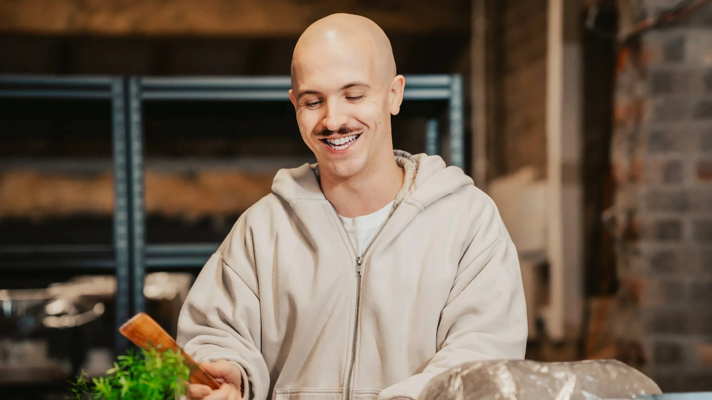Artist Todd Gilbey, co-founder of Northamptonshire luxury ceramics studio FOUND, smiling while shaping clay with a wooden tool against a rustic industrial background