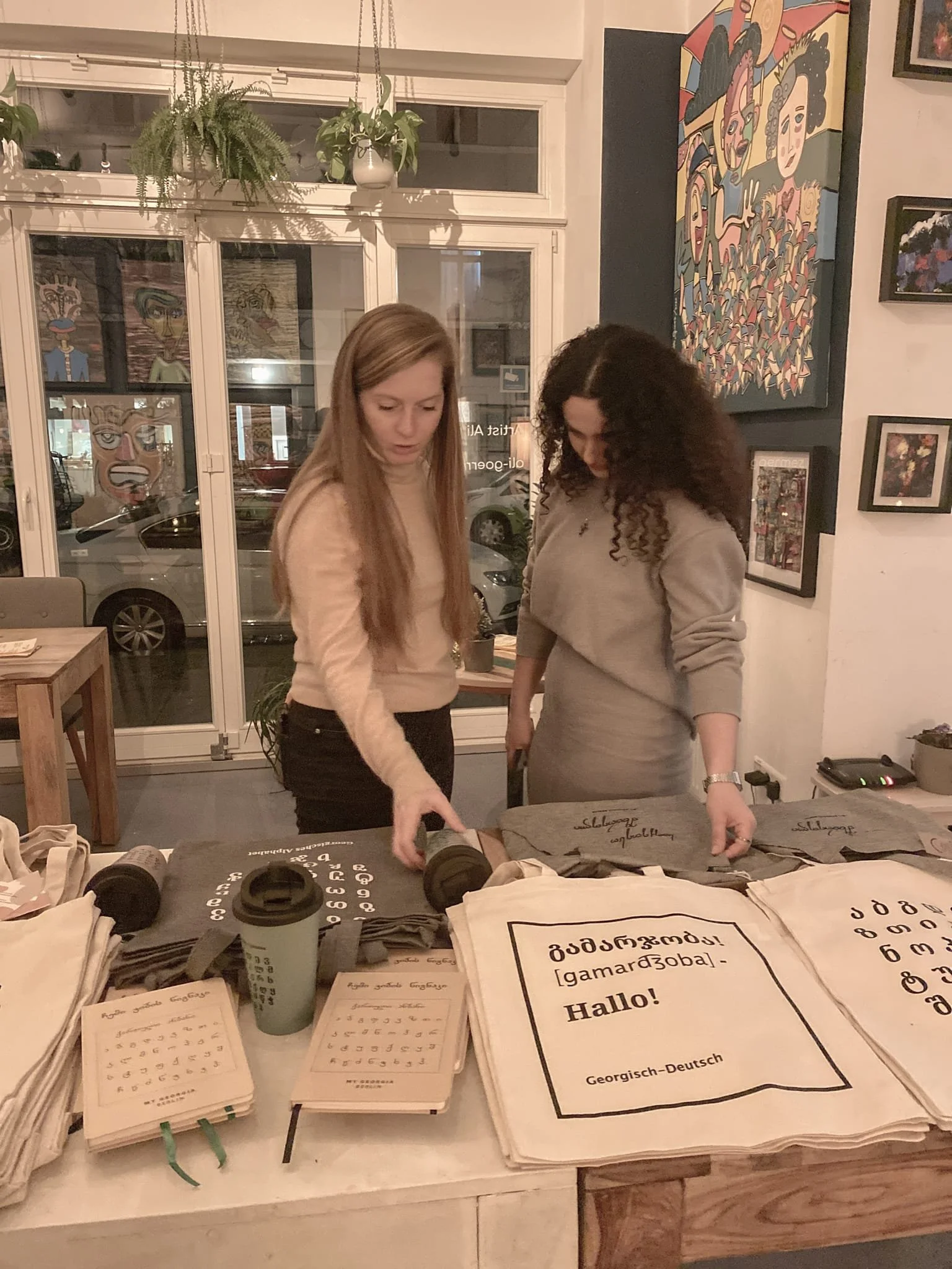 Zwei Frauen schauen sich handgeschriebene Materialien und Stofftaschen auf einem Tisch in einer Kunstgalerie oder einem Gemeinschaftsraum mit Gemälden an der Wand an.