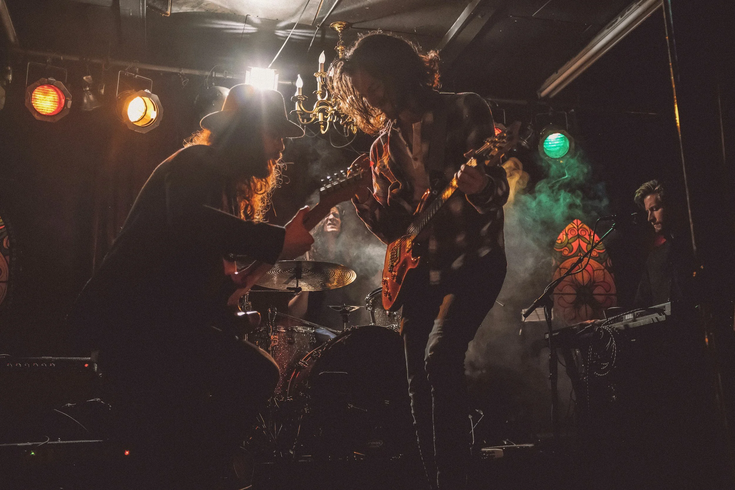 A group of four musicians performing on stage. The focus is on two guitar players, one with curly hair and the other with wavy hair, facing each other. A drummer and keyboardist are in the background. The stage is dimly lit with colorful lights and smoke, creating a moody atmosphere.