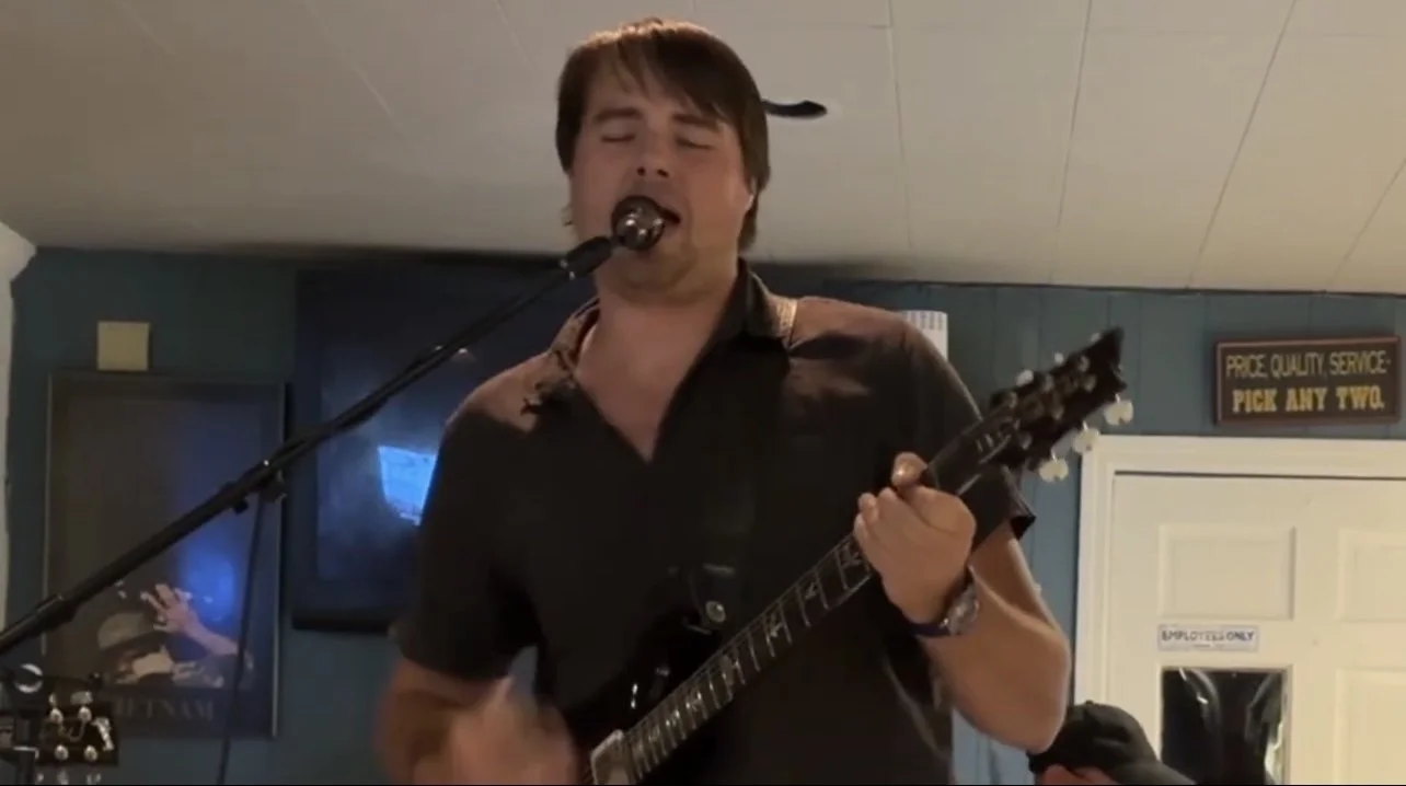 Man singing into a microphone while playing an electric guitar indoors.