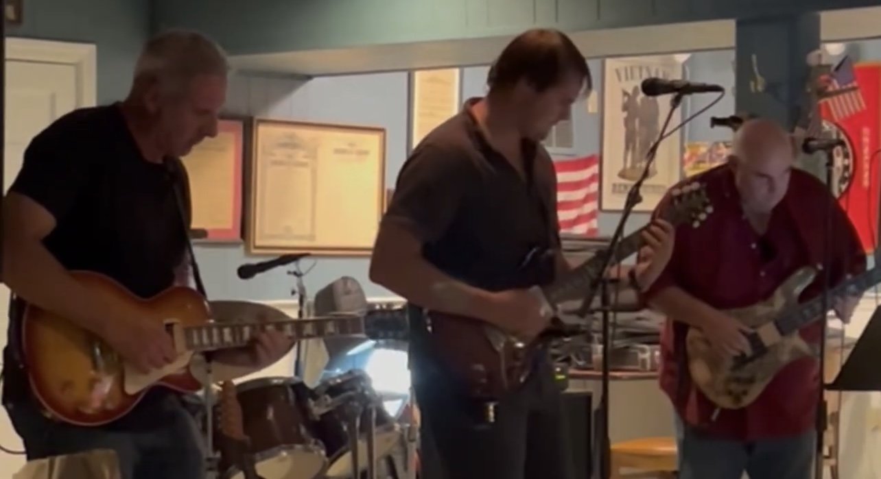 Three men playing musical instruments in a bar or restaurant, one with an electric guitar, one with a bass guitar, and one with a mic stand, during a live performance.