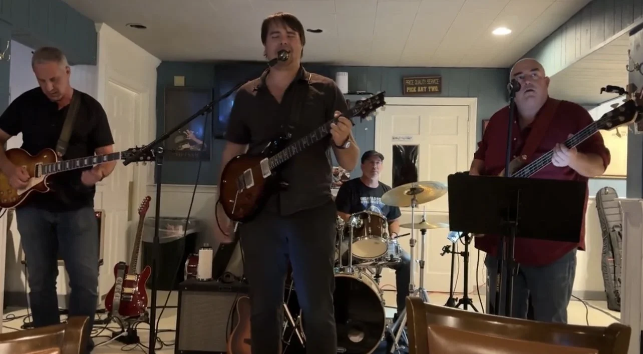 A band of four musicians performing indoors, with two guitarists, a drummer, and a bassist.