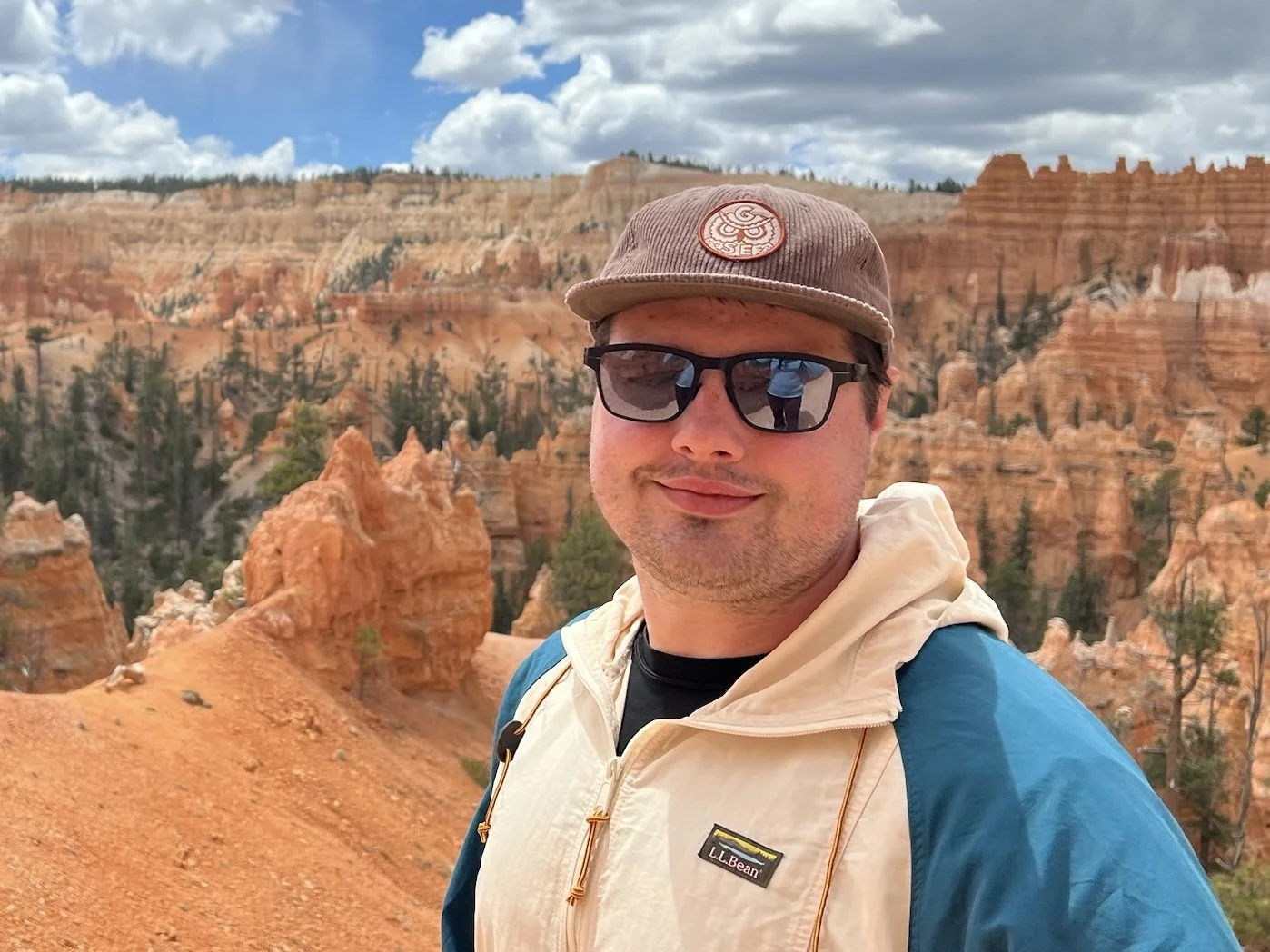 A man wearing sunglasses, a beige L.L. Bean jacket, and a brown cap stands in front of colorful rock formations and trees in a canyon with a partly cloudy sky.
