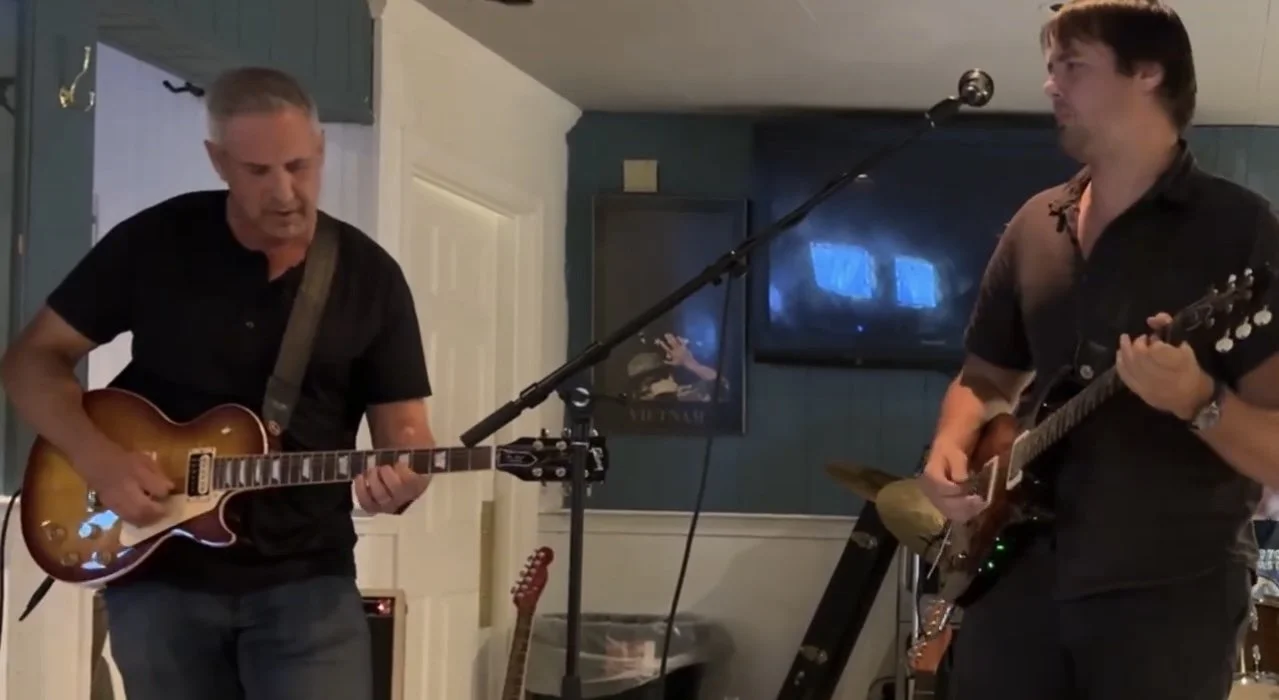 Two men playing electric guitars in a room with a TV and a picture on the wall.