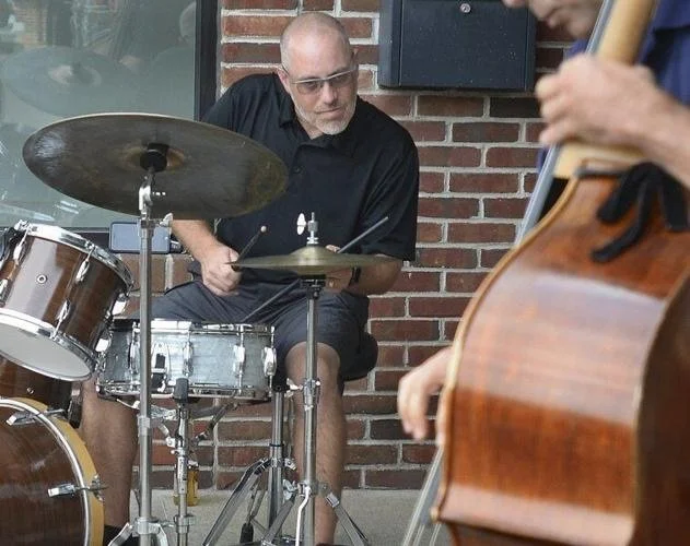 A man playing drums and another playing guitar outdoors in front of a brick wall.