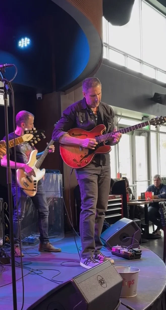 Man playing electric guitar on stage, two other musicians playing bass guitars nearby, with audience members seated at tables nearby.