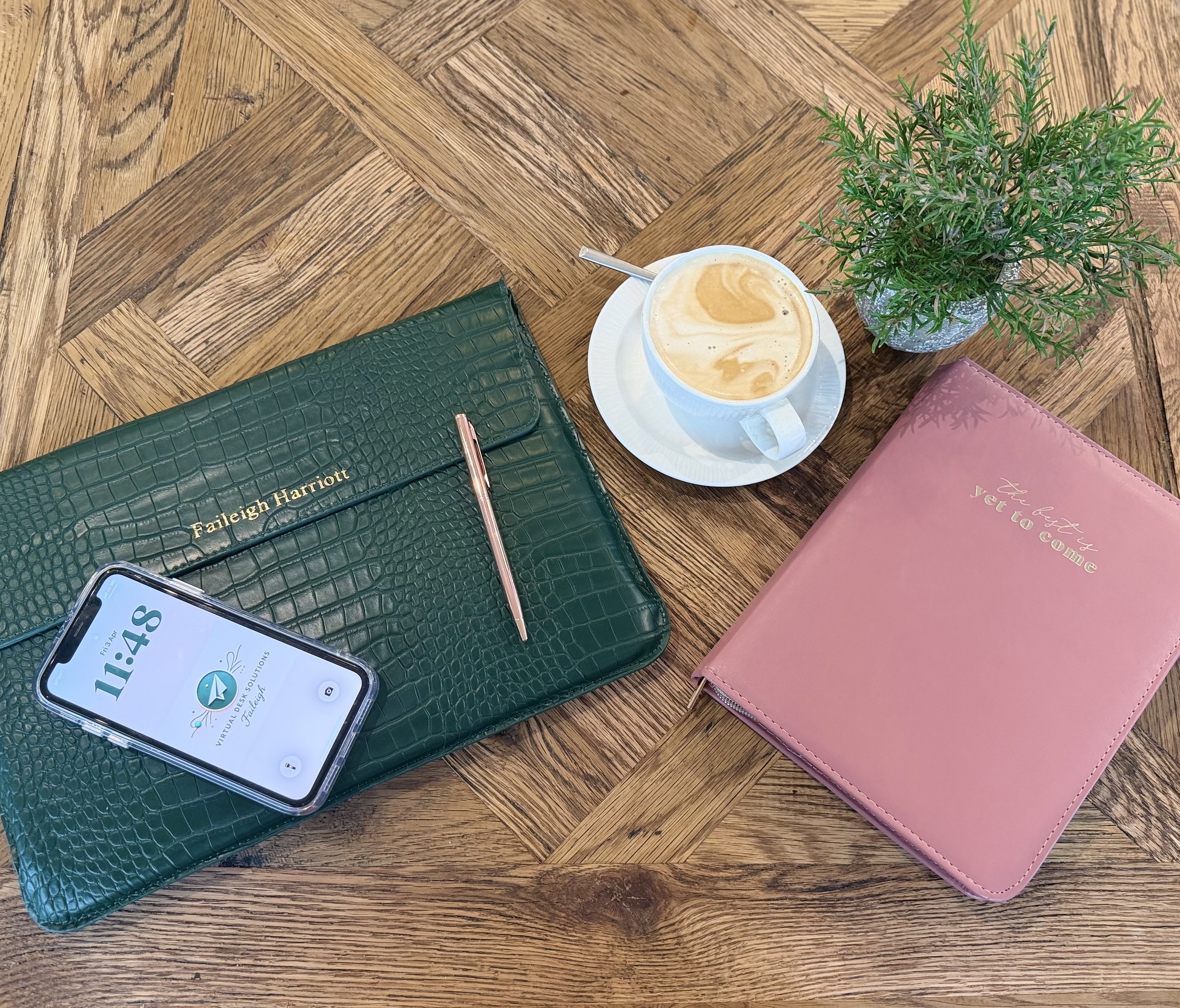 A top view of a wooden table with a potted plant, a cup of coffee with a spoon, a green wallet, a pink notebook, and a smartphone displaying the time 11:44.