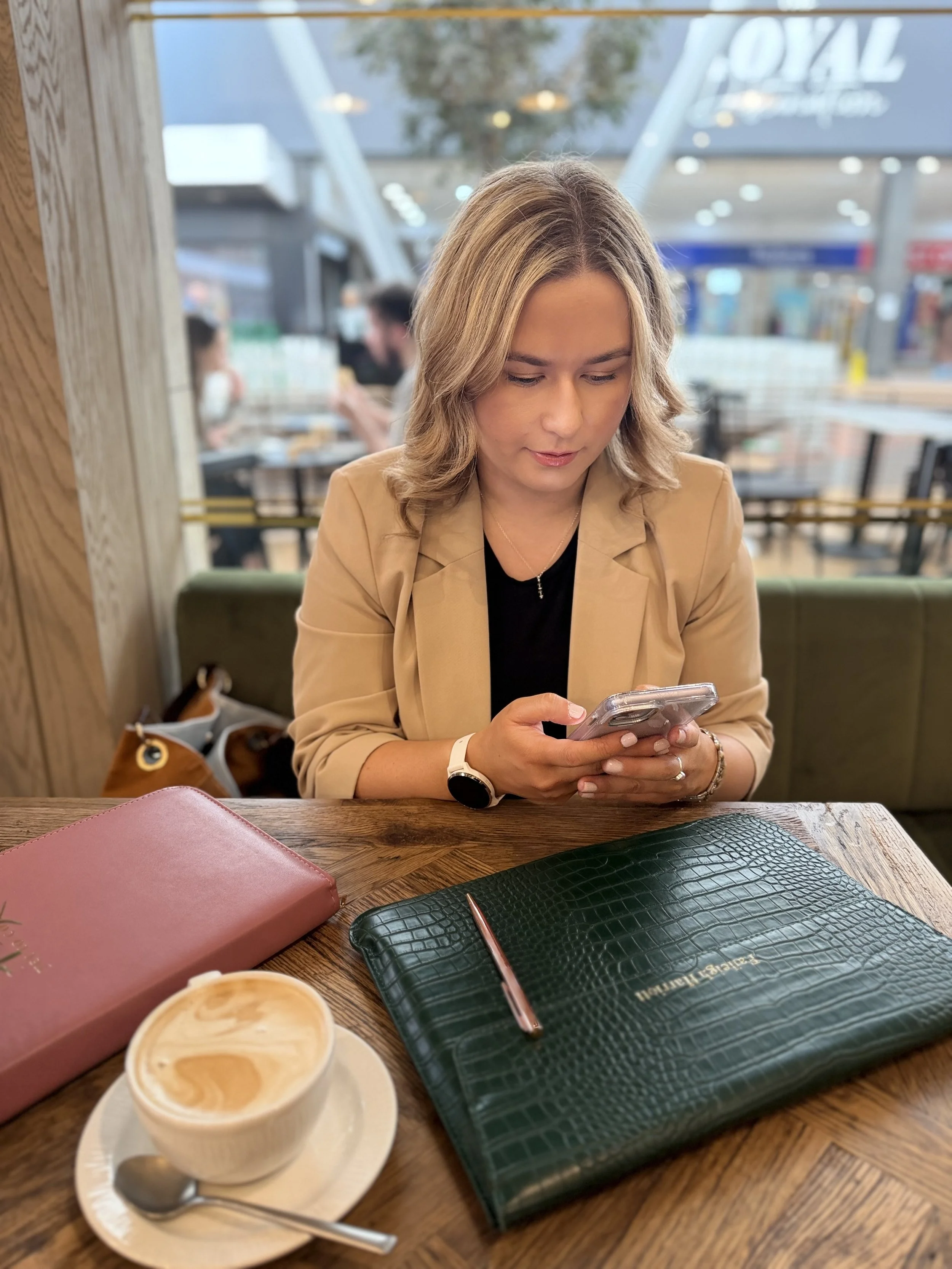 Woman with blonde hair sitting at a table in a cafe, looking at her phone. On the table are a cup of coffee, a pink leather wallet, a black textured notebook with a rose gold pen, and another padded item. The background shows large windows with people sitting and talking.