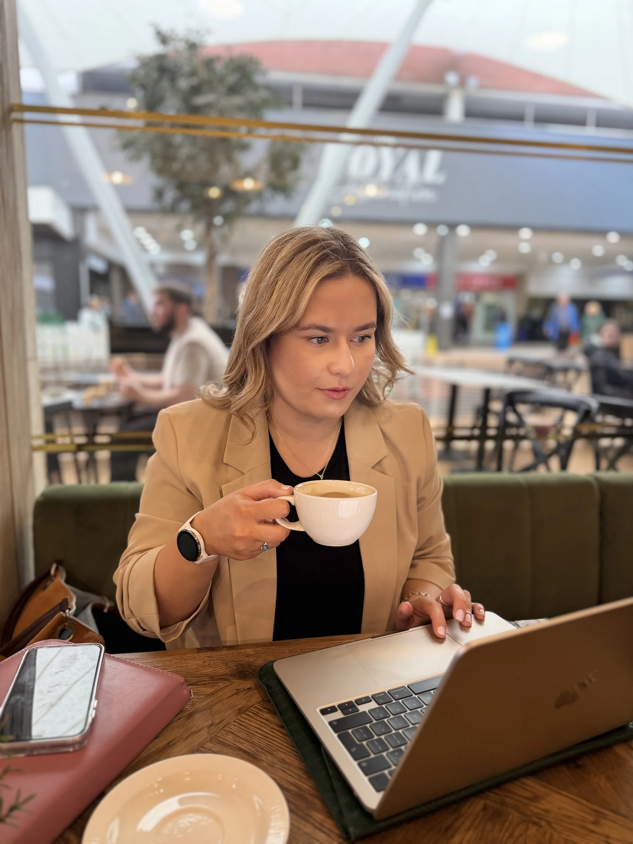 A woman with blonde hair seated at a table in a coffee shop, holding a white cup of coffee, with a laptop, smartphone, and pink bag on the table, and a window behind her showing a mall or airport.