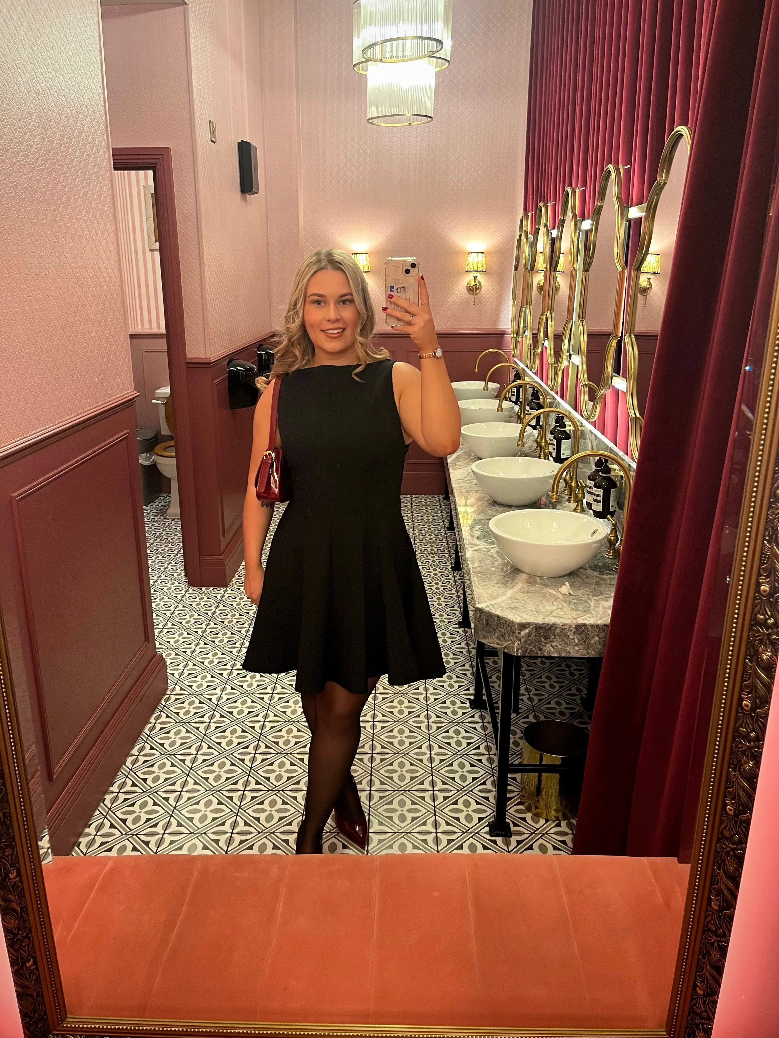 A young woman with blonde hair takes a selfies in front of a mirror in a bathroom with pink walls, patterned tiles, and velvet curtains; she is dressed in a black sleeveless dress, black stockings, and red shoes, holding a red handbag.