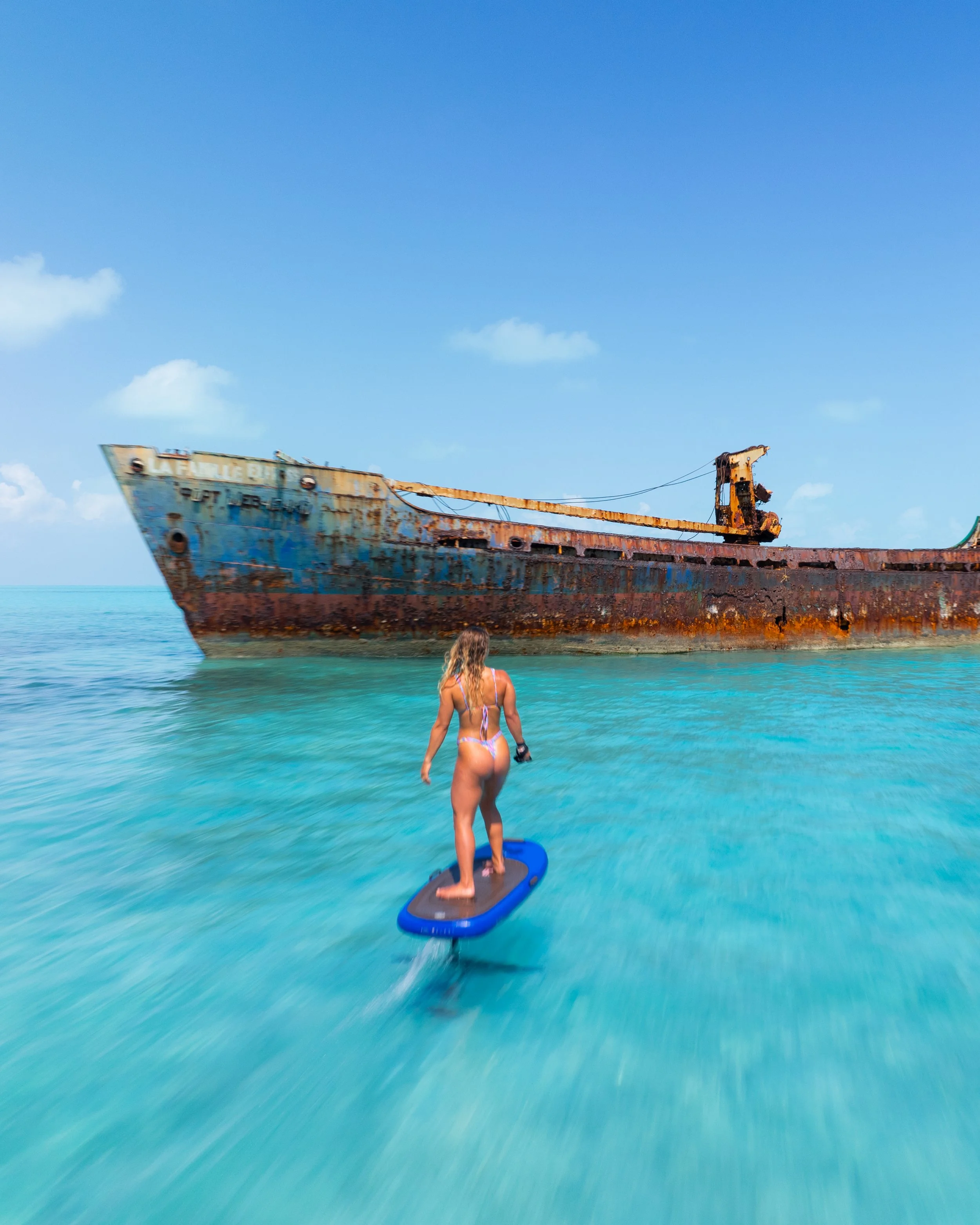 A woman in a bikini efoiling in clear blue water near a rusted, abandoned shipwreck under a partly cloudy sky. Located in Gracebay, Providenciales, Turks & Caicos.