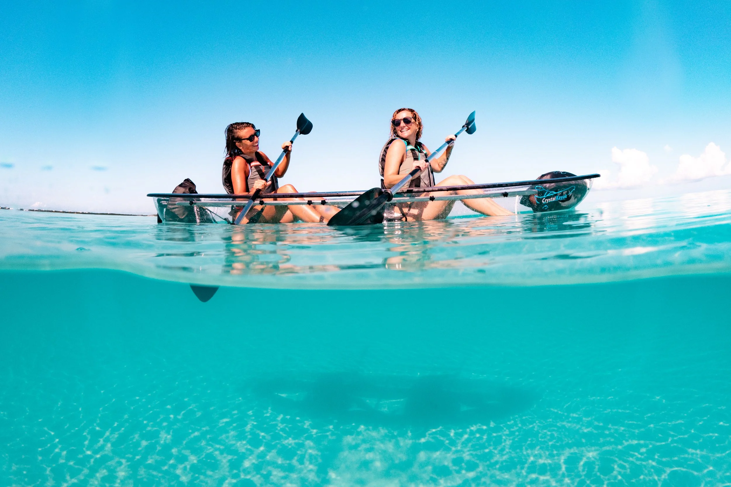 Two happy women in clear bottom kayak in the ocean on a sunny day, wearing life jackets and sunglasses, with clear blue water and sky. Located in Turks & Caicos.