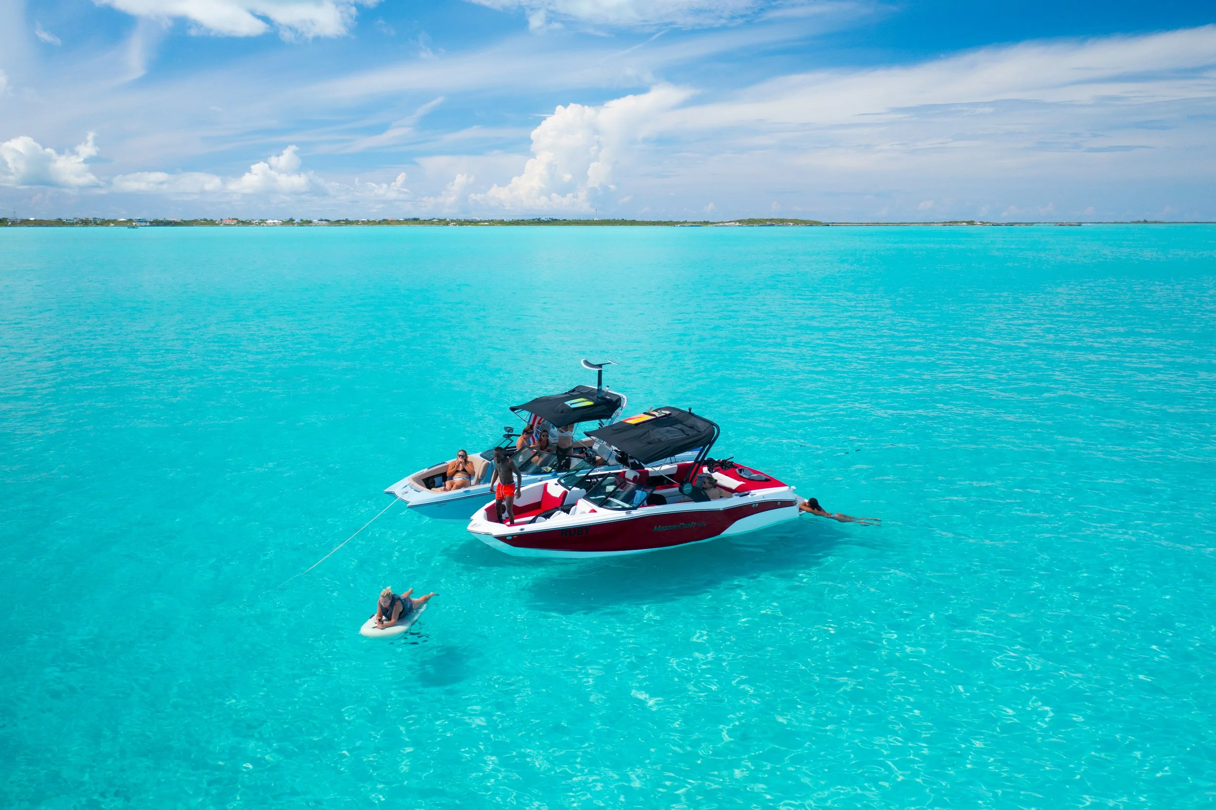 Two wake boats with black canopies on turquoise water, with people relaxing on both boats and one person swimming near the watercrafts. Clear sky with some clouds in the background. Perfect for wakesurfing, wakeboarding, waterskiing, tubing. Located