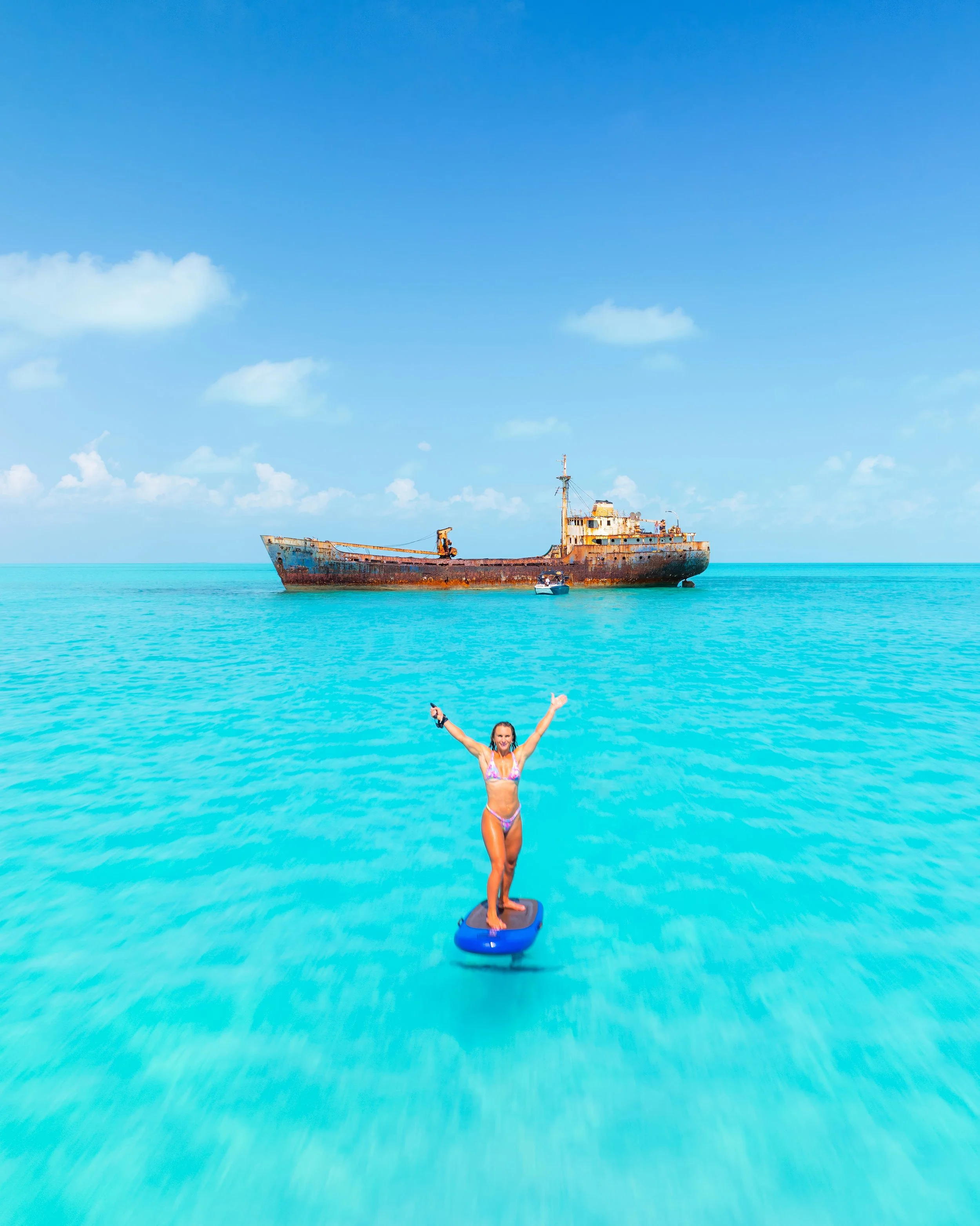 A woman in a bikini standing on an efoil with arms raised, floating on turquoise water with a rusty shipwreck in the background under a partly cloudy sky. Perfect for wakesurfing, wakeboarding, waterskiing, tubing. Located in Gracebay, Providenciales